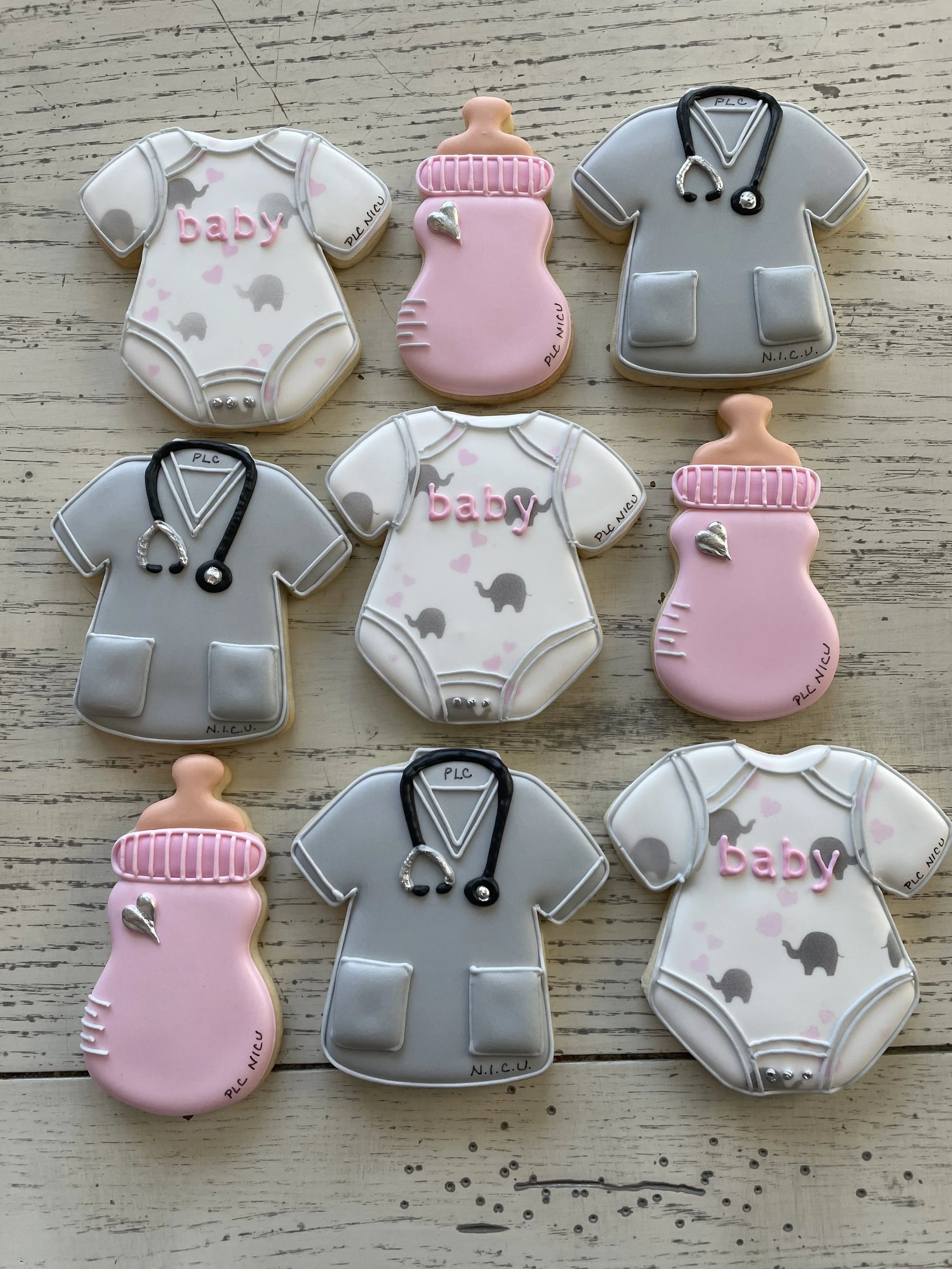Cookies decorated as baby onesies, baby bottles, and medical scrubs with stethoscope details, arranged on a wooden surface.