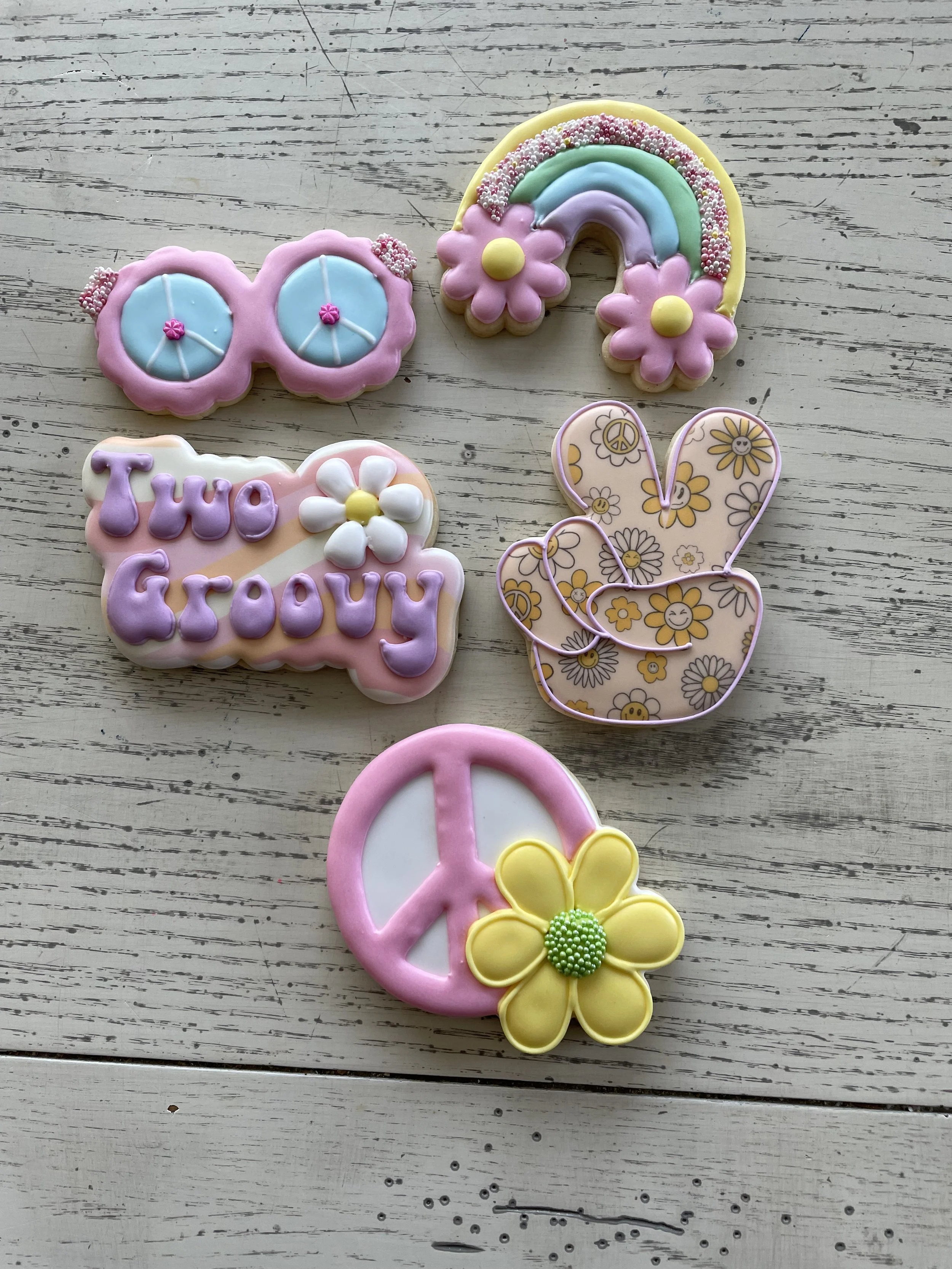 Colorful decorated cookies including sunglasses, a rainbow, a speech bubble with flowers, a peace sign with a flower, and a bunny shape, all on a white wooden surface.