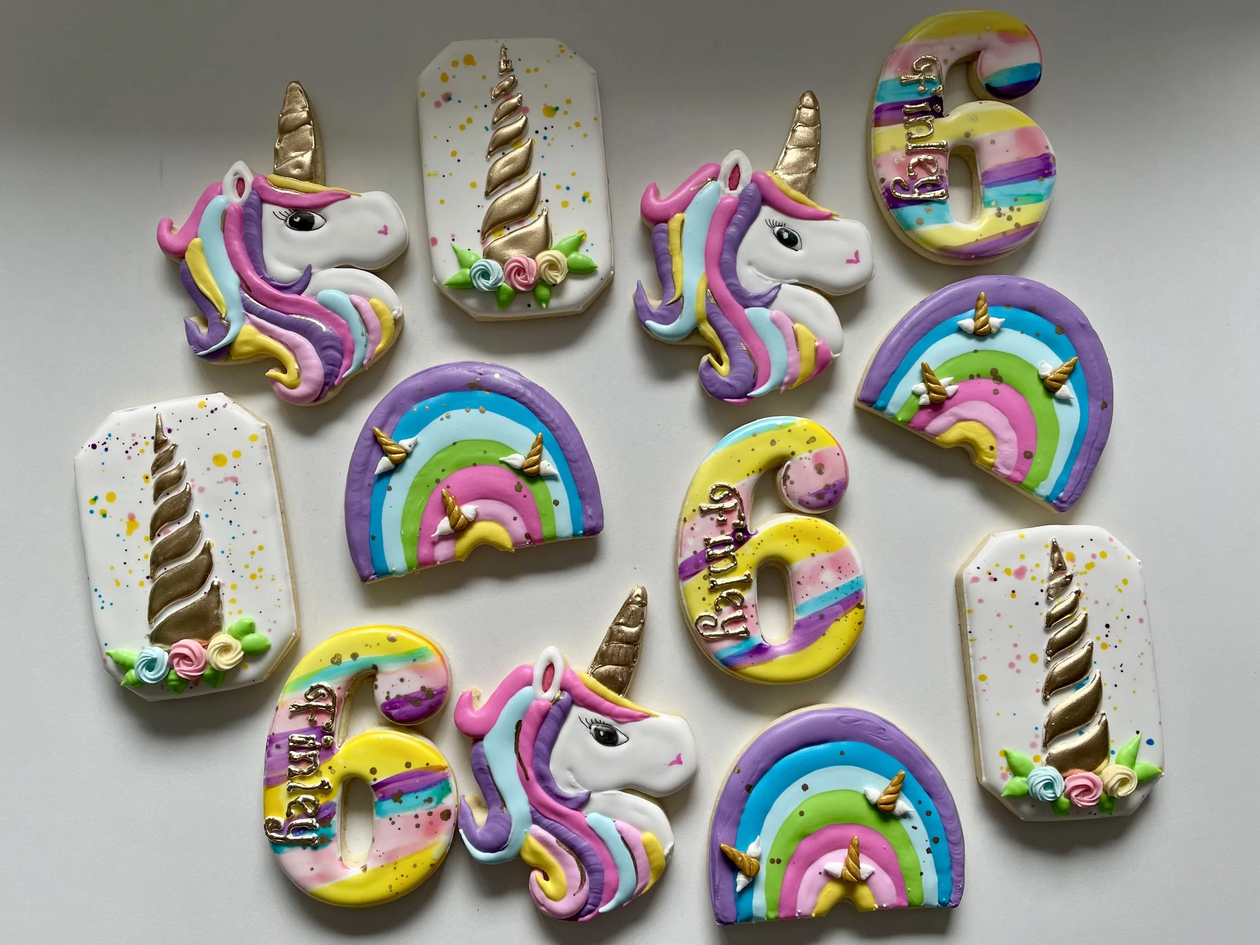Decorative cookies shaped and decorated as unicorns, rainbows, the number 6, and candles with 'Happy Birthday' written, arranged on a white surface.