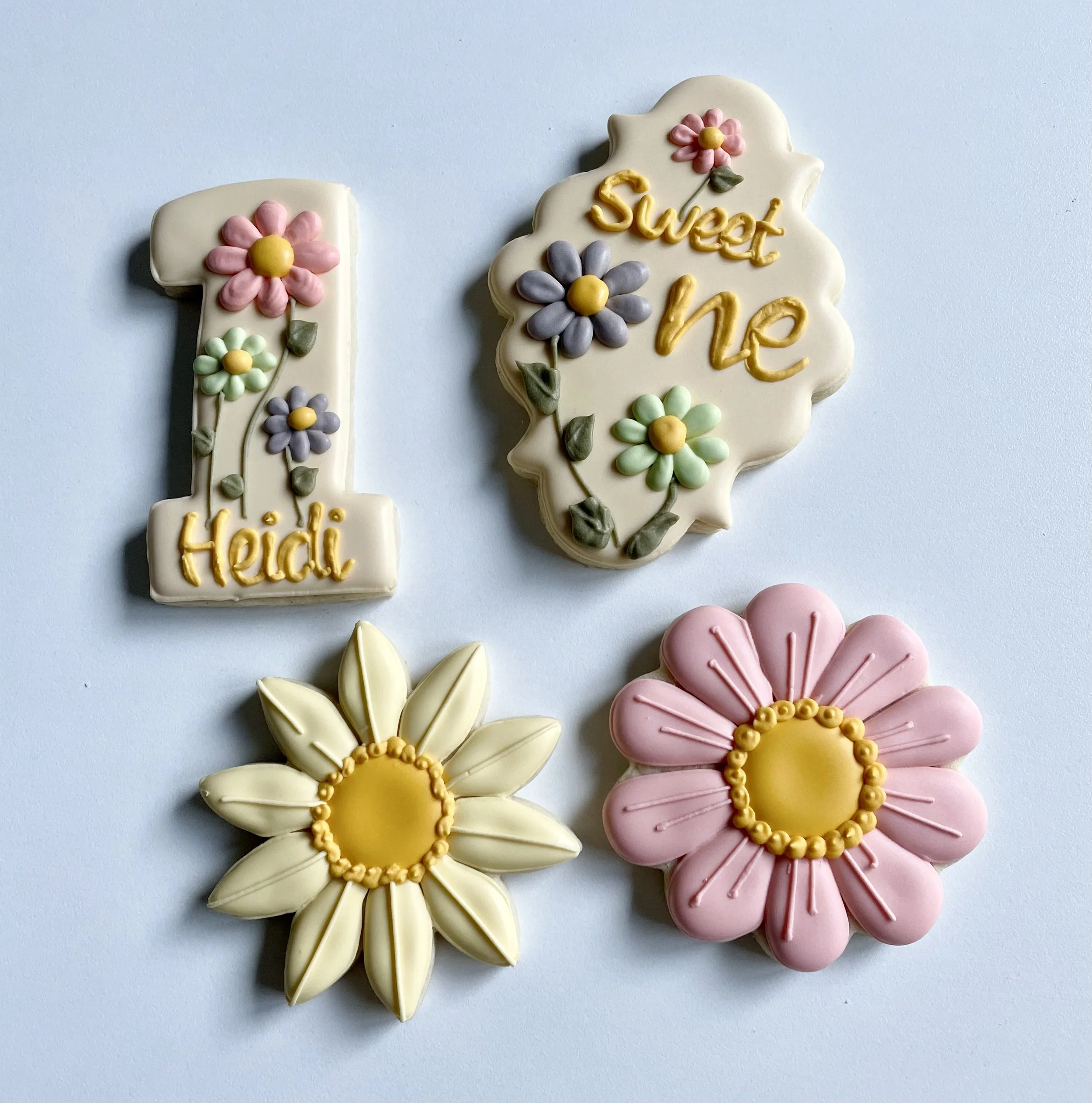 Decorated cookies with floral designs and personalized message, including a large number one cookie with pink, green, and purple flowers and the name Heidi, a scalloped edge cookie with the message "Sweet one" and floral decorations, and two flower-s