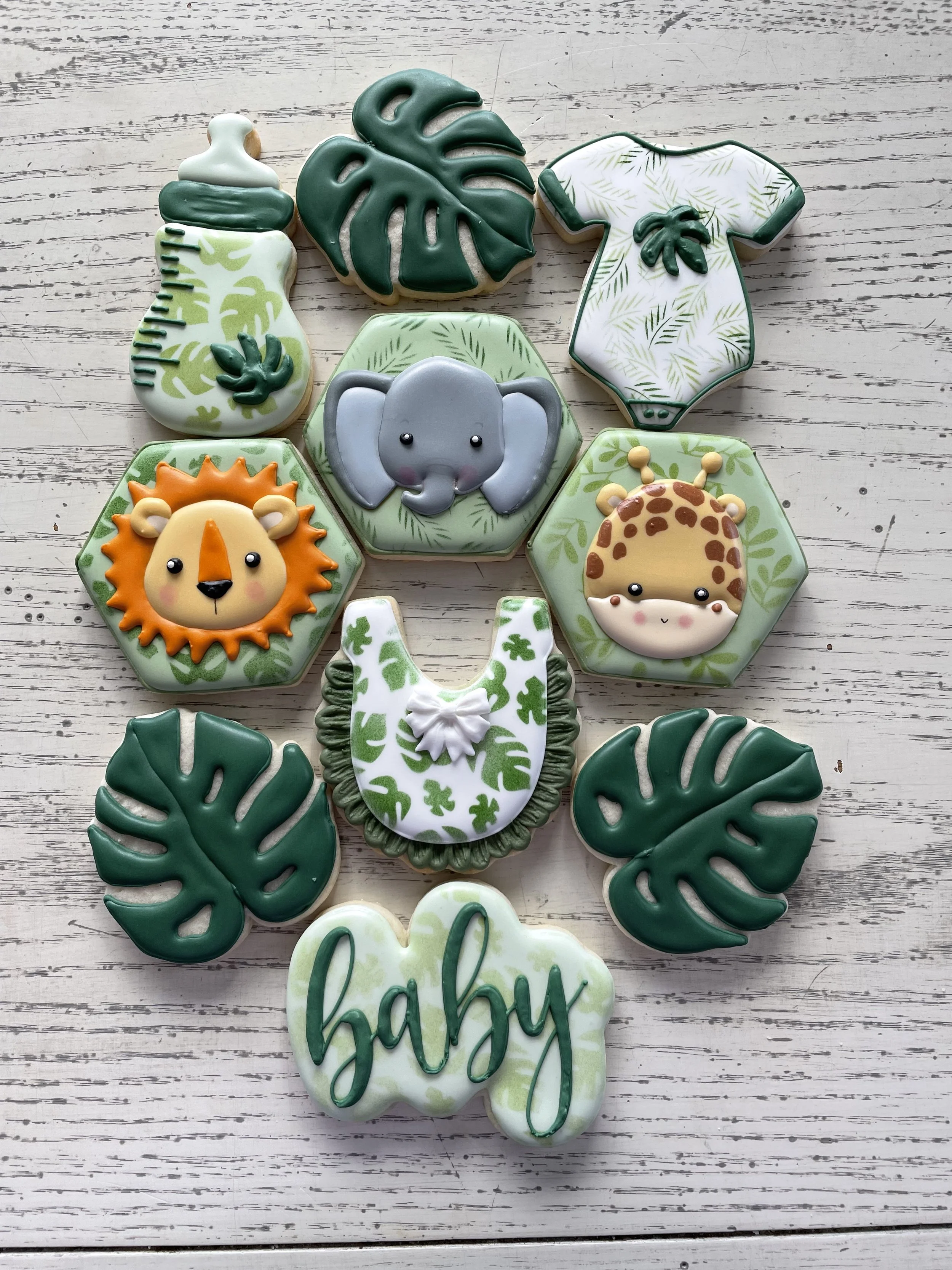 Collection of jungle animal-themed cookies including a baby bottle, monstera leaves, a onesie, a lion, an elephant, a giraffe, a bib, and the word "baby" on a light wood background.
