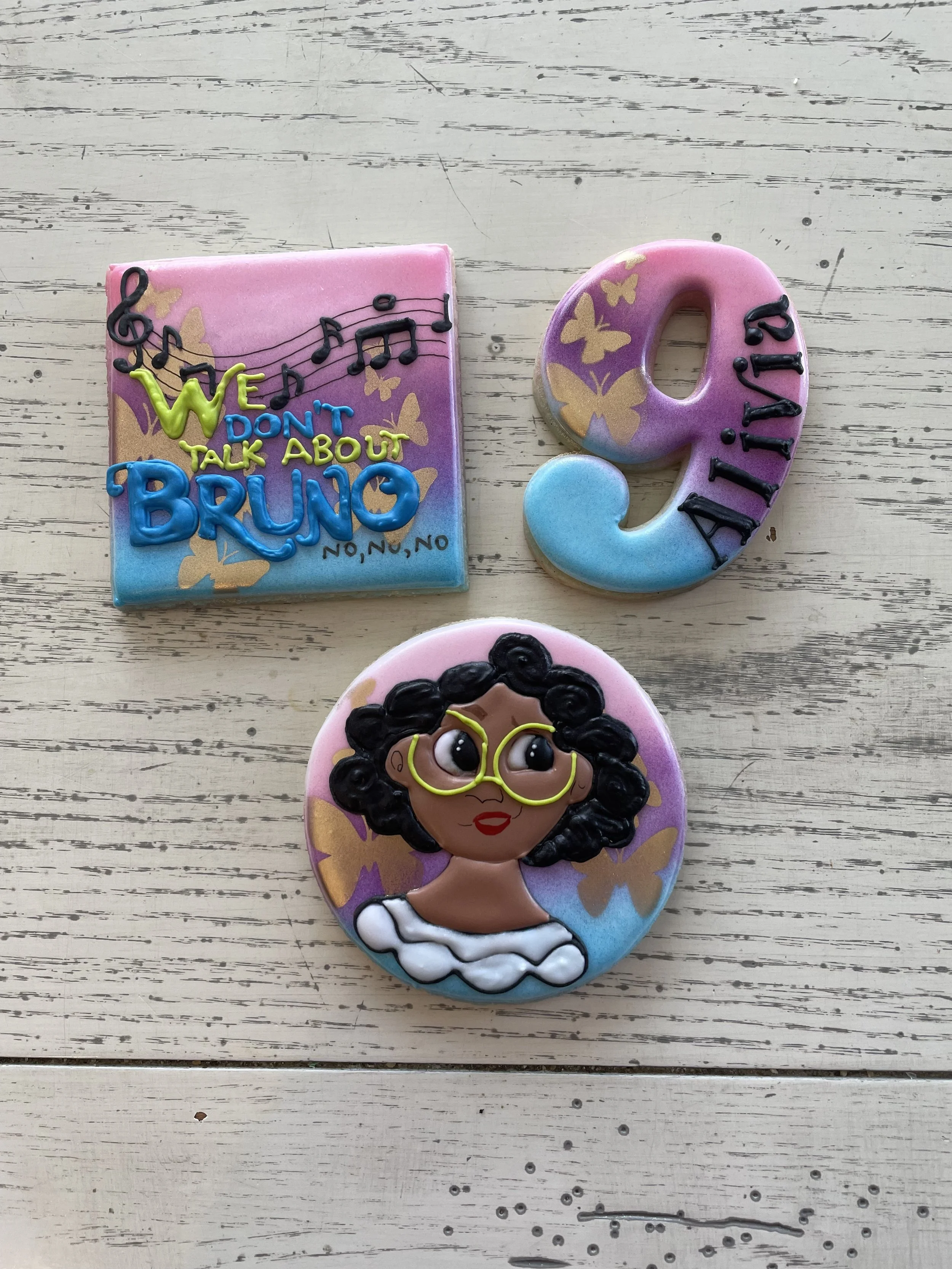 Decorative cookies with colorful designs and messages, including a square cookie with music notes and the words 'We don't talk about Bruno,' a number 9 cookie with a butterfly design, and a round cookie featuring an illustration of a girl with curly