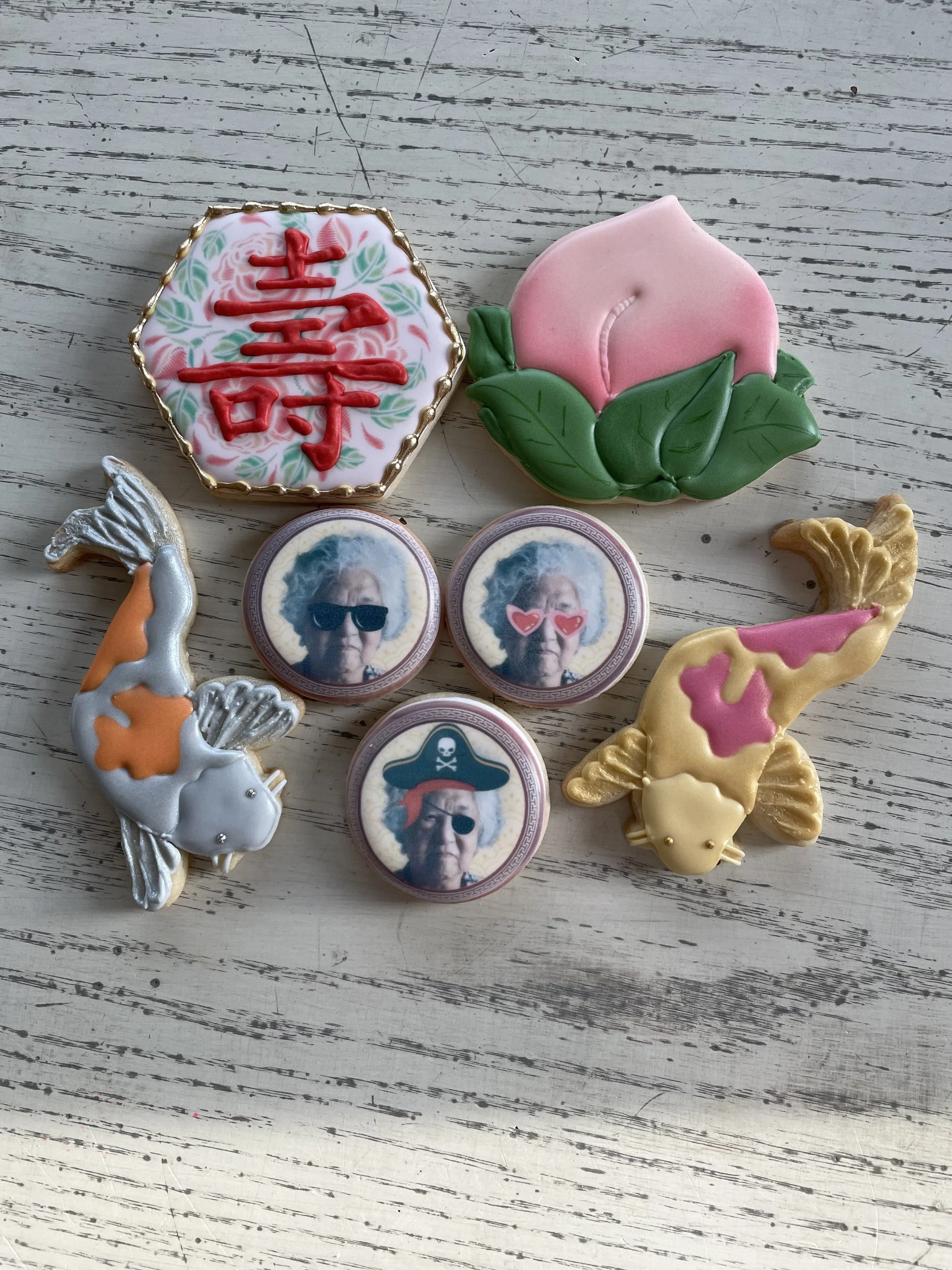 Decorative cookies with themes of age, love, and humor, featuring a peach blossom, Chinese characters, elderly women in sunglasses with added accessories, and cartoon fish with gold accents, all arranged on a white wooden surface.