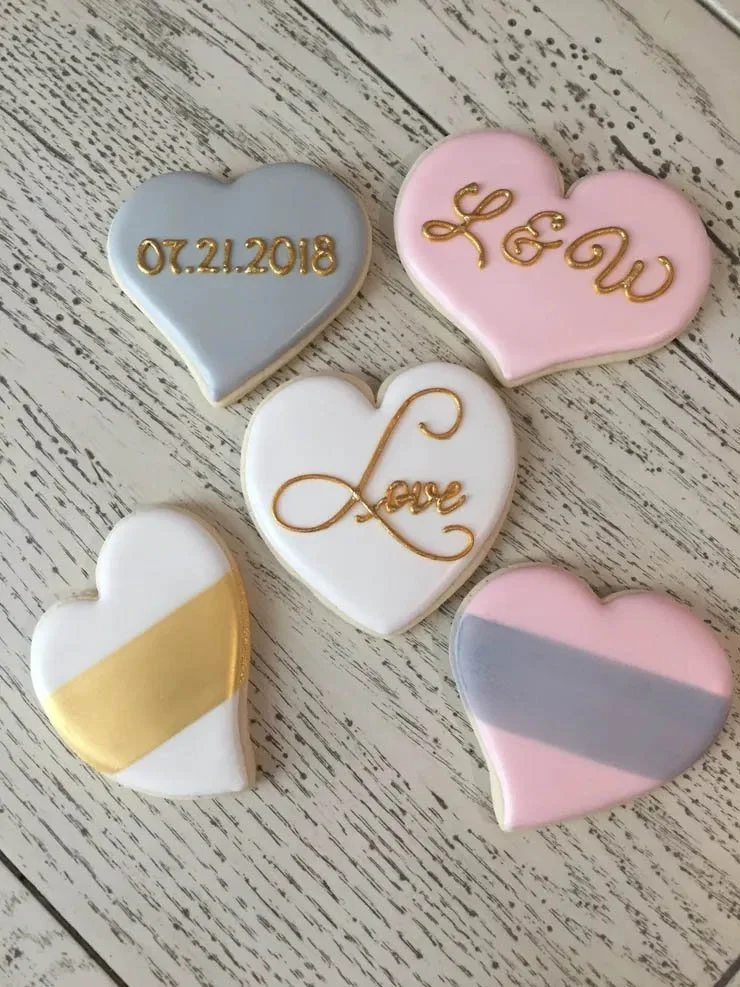 Five decorated heart-shaped cookies with pastel-colored icing and gold lettering or accents, arranged on a white wooden surface.