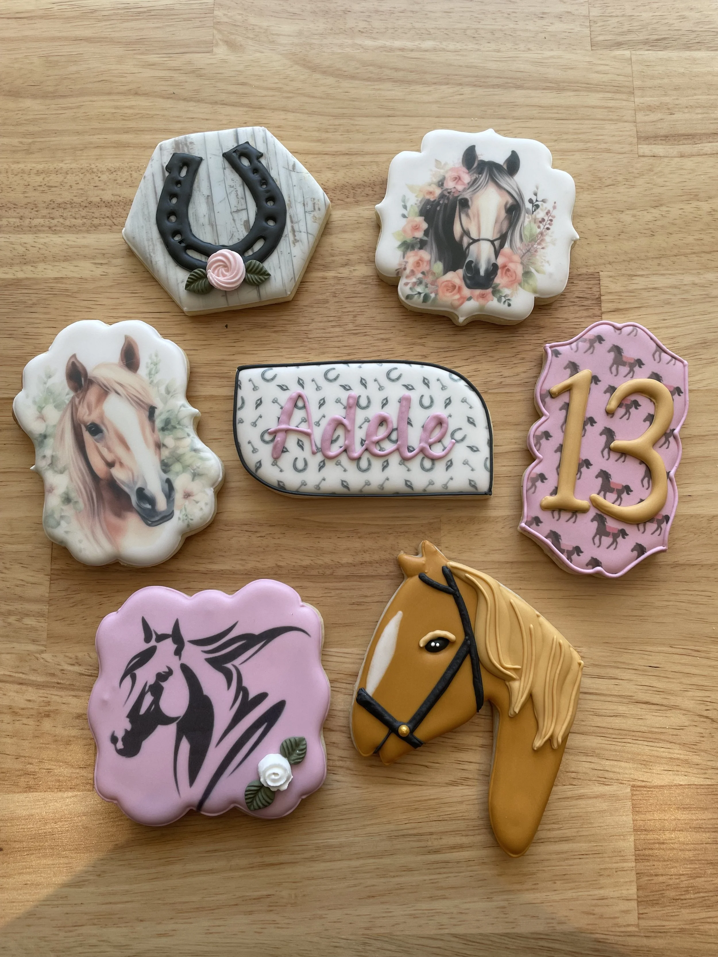 Collection of decorated horse-themed cookies on a wooden surface, featuring horseshoes, horse heads, a sign with the name 'Adele', the number 13, and a horse's head.