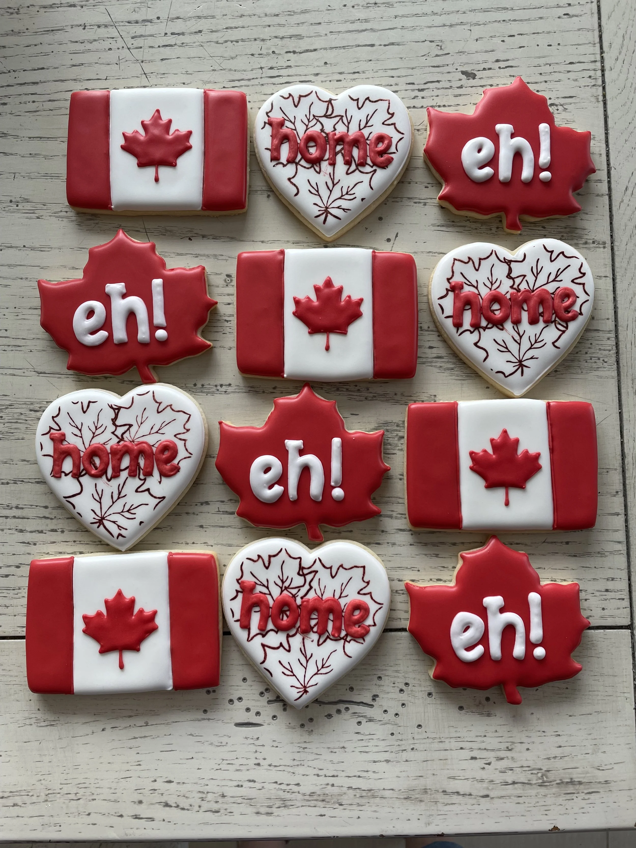 Decorative cookies with Canadian flag, maple leaf symbols, and the words "home" and "eh!" in red and white colors on a light wooden surface.