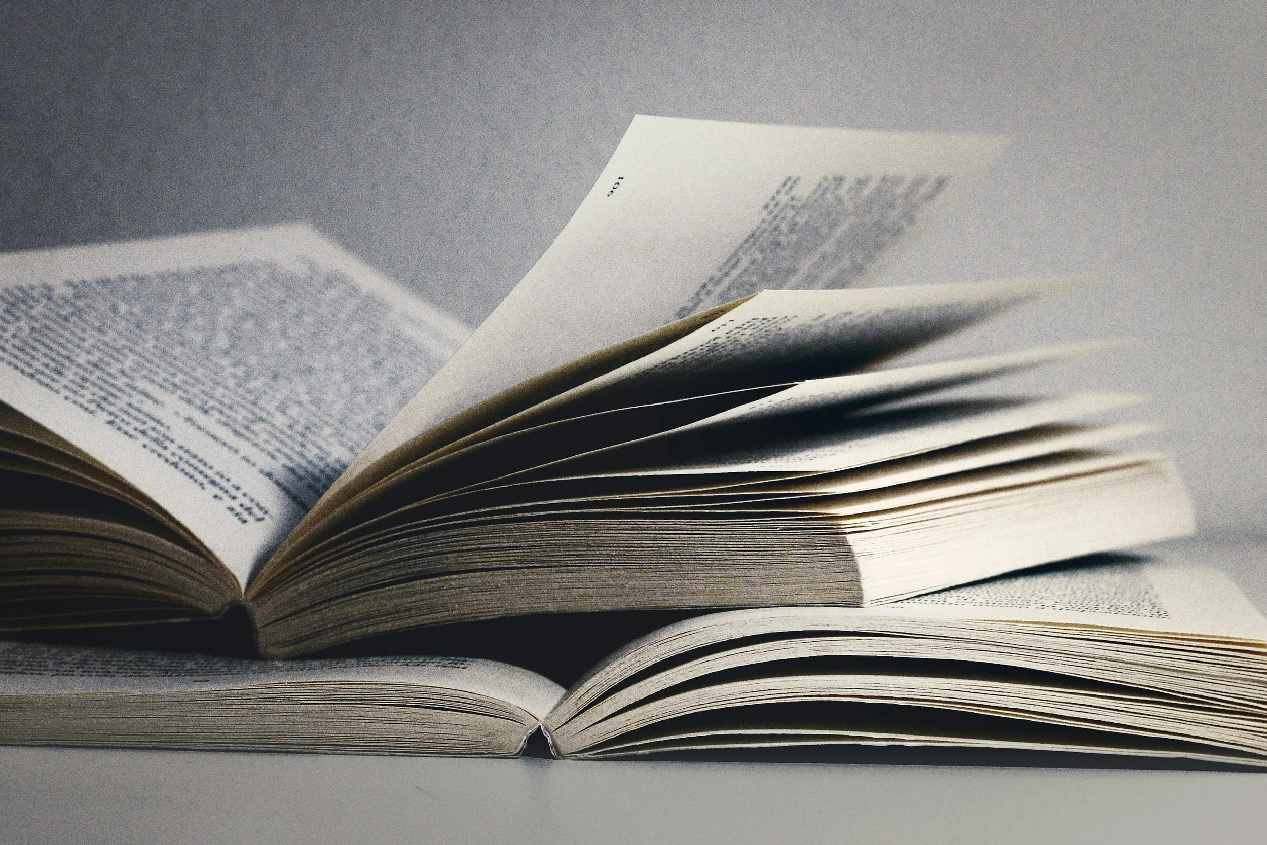Two open books stacked together, their cream-colored pages fanning upward against a gray background.