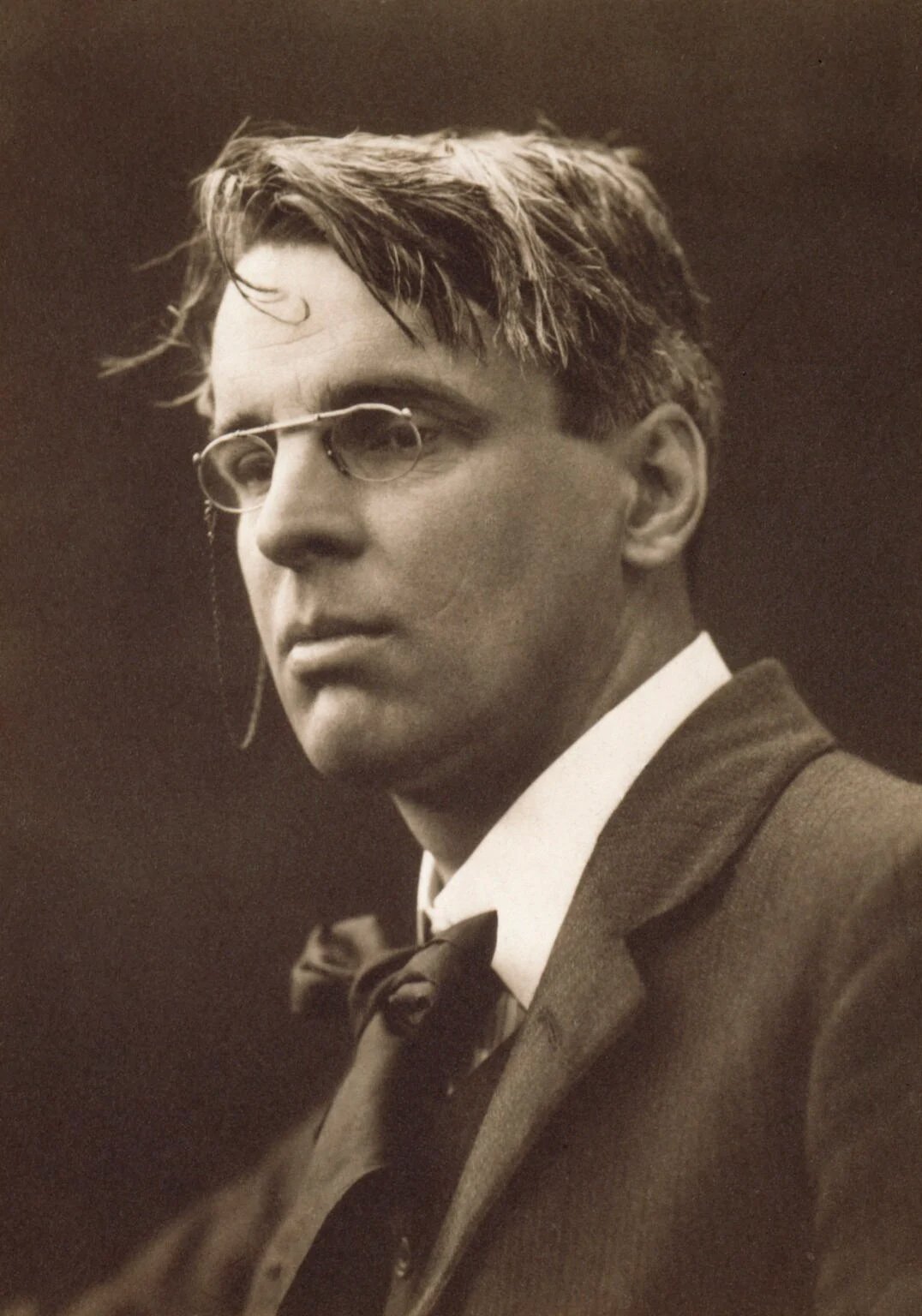 Sepia portrait of W. B. Yeats wearing glasses and a bow tie, his face turned slightly to the left.