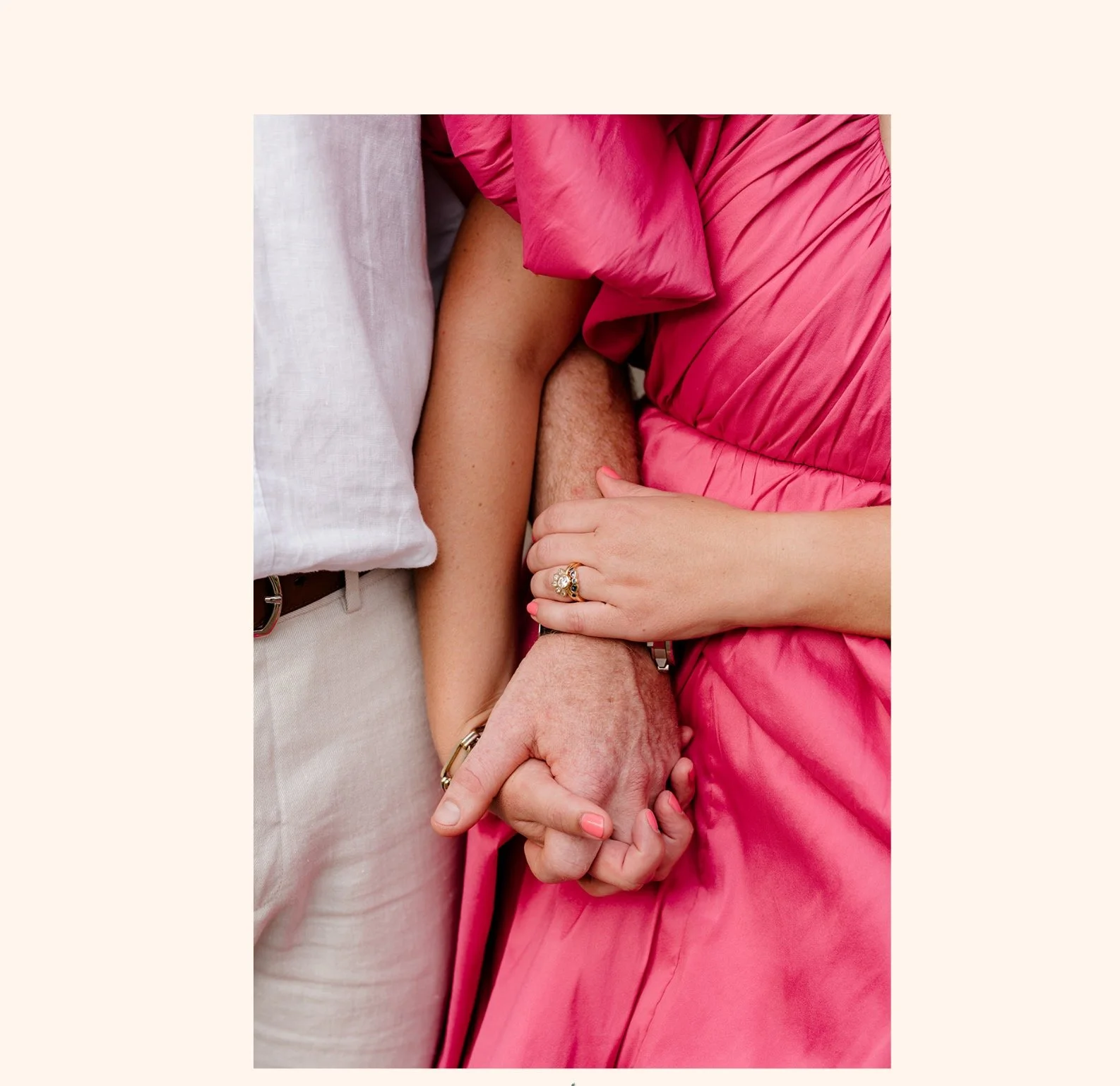 Close-up of a couple holding hands, with the woman wearing a pink dress and an engagement ring, and the man wearing a white shirt.
