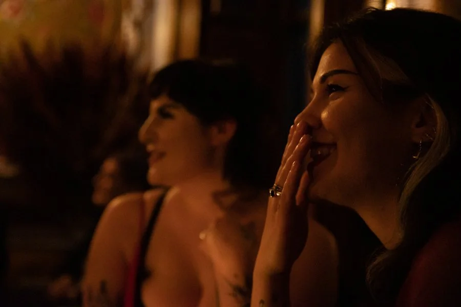 Two women laughing and smiling in a dimly lit indoor setting, one woman covering her mouth with her hand.