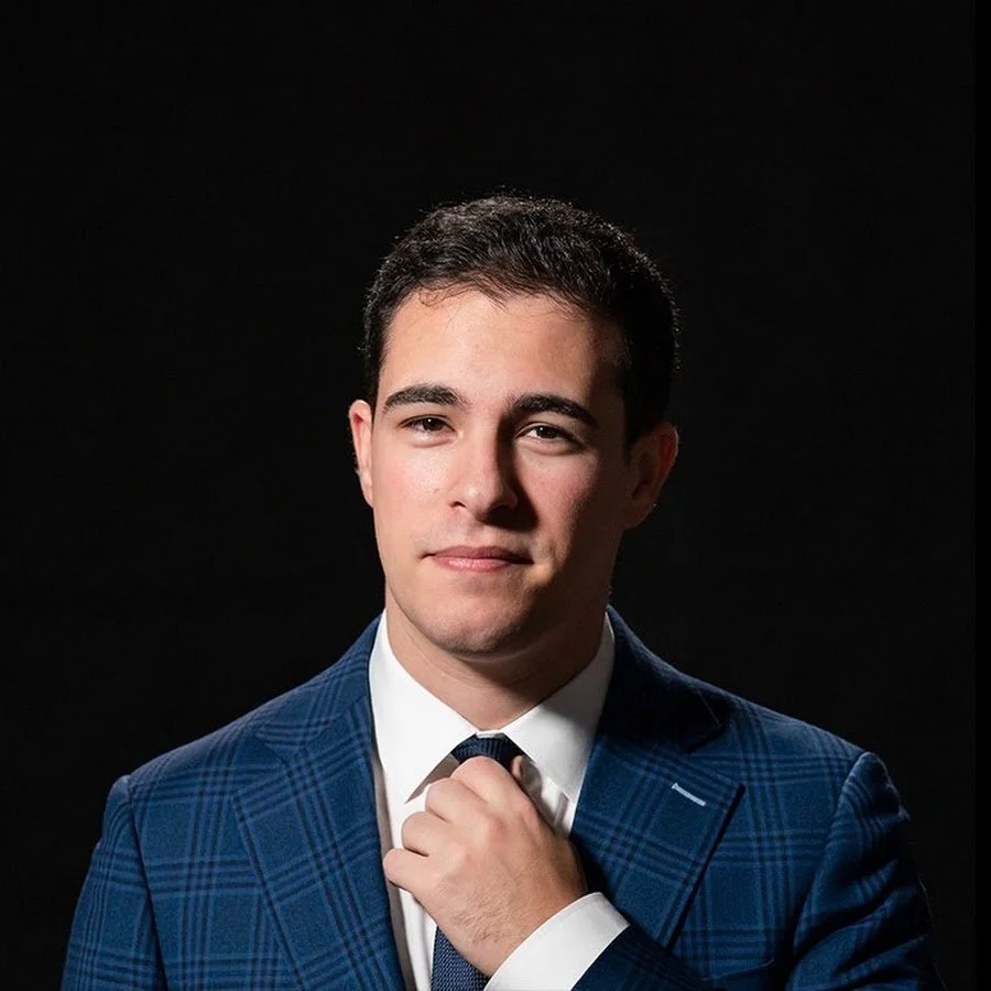 A young man with short dark hair, wearing a blue checkered suit, white shirt, and dark tie, adjusting his tie against a black background.
