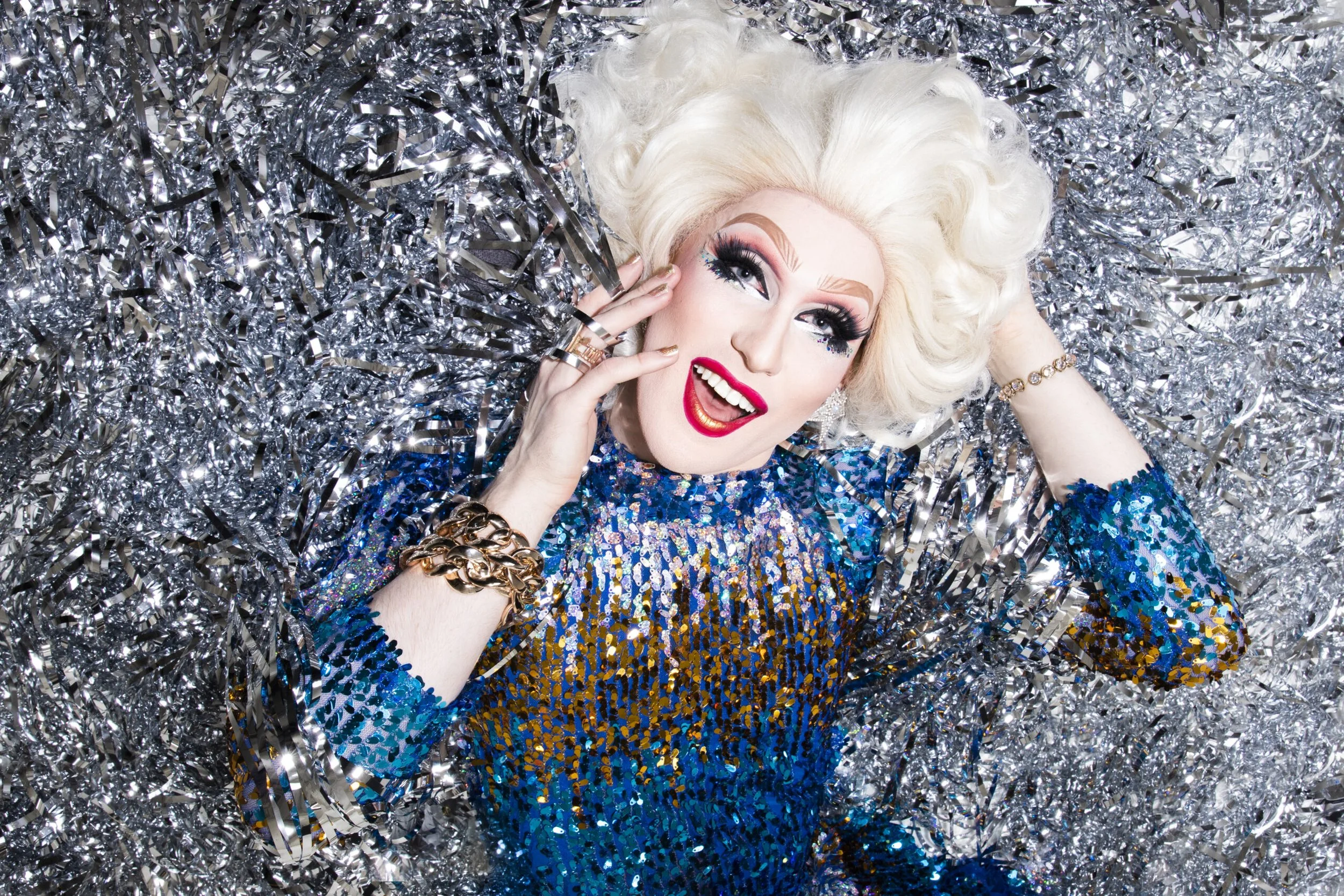 A drag queen with platinum blonde hair, dramatic makeup, and a blue sequin dress lying on silver confetti and tinsel, smiling and holding one eye open.