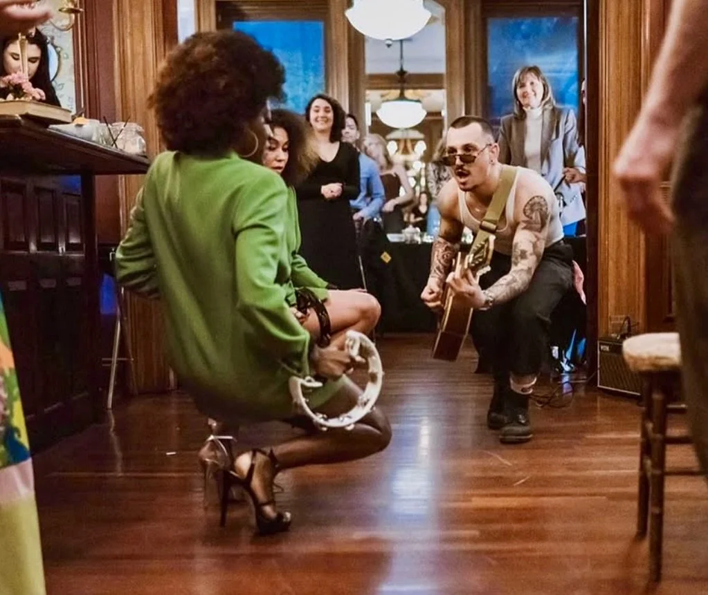 A man with tattoos, sunglasses, and a tank top is playing guitar and singing to a woman in a green blazer who is holding a tambourine, while a group of people watch and smile in a warmly lit room with wood-paneled walls and a chandelier.