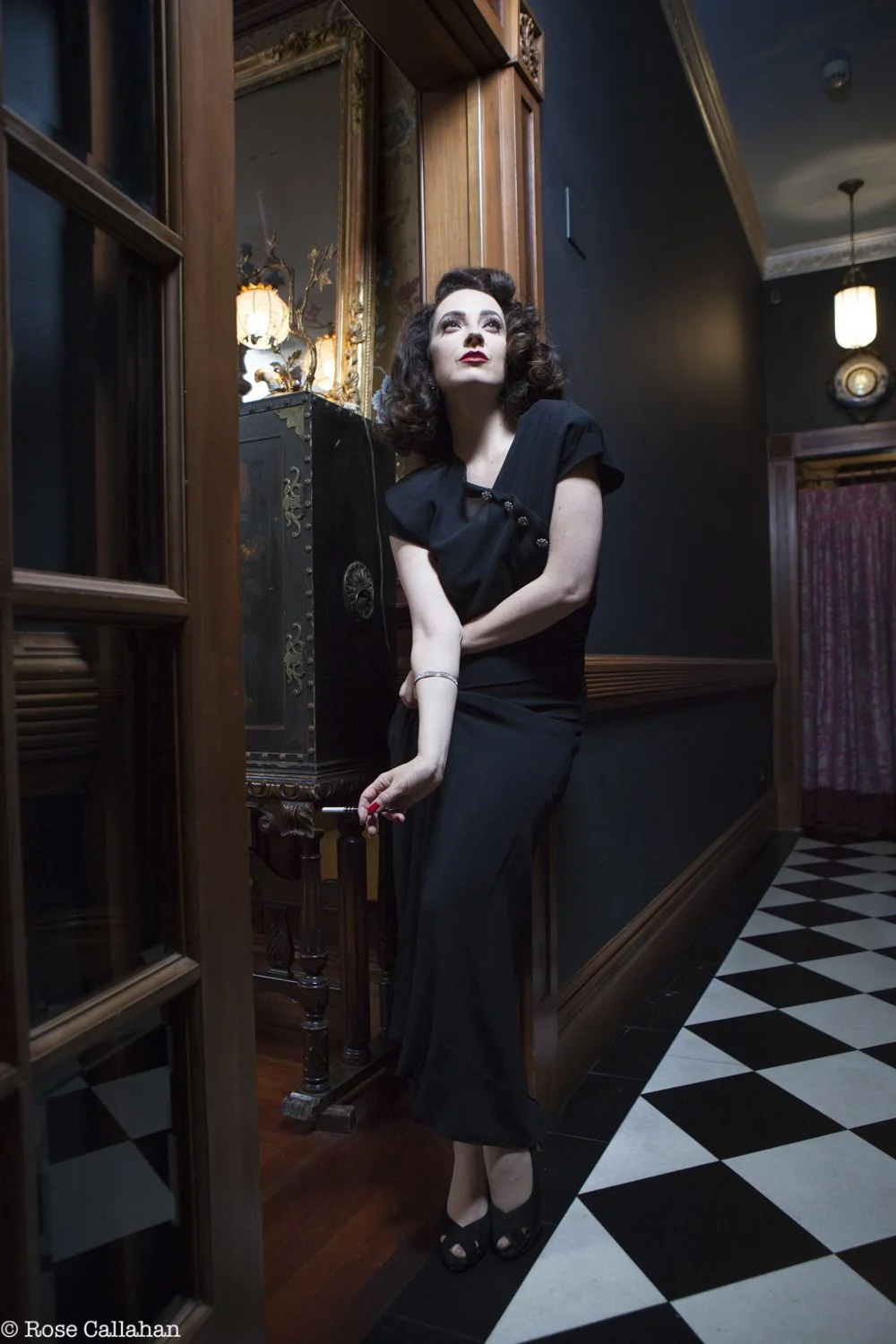 A woman in a black dress with vintage style, holding a cigarette, standing in a dimly lit room with dark walls, wooden accents, a mirror, and a checkered black and white floor.