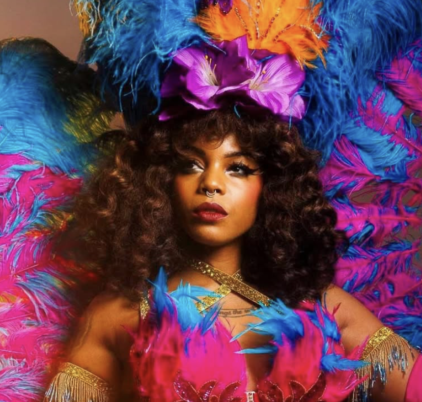 Woman wearing a colorful feather headdress with pink, blue, purple, and orange feathers, dressed in a vibrant costume with feathers and glitter, standing against a neutral background.