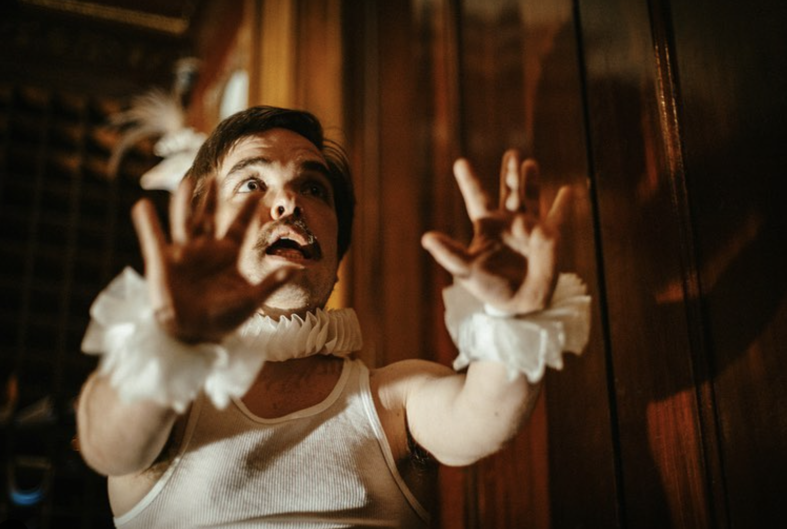 A man dressed in a white tank top and ruffled cuffs, with a surprised or frightened expression, reaching out with both hands towards a wooden surface or wall.