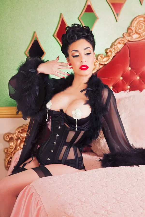 A woman with dark, styled hair and bright red lipstick posing on a bed with an ornate gold headboard, wearing black lingerie and a black sheer robe with feather trim.