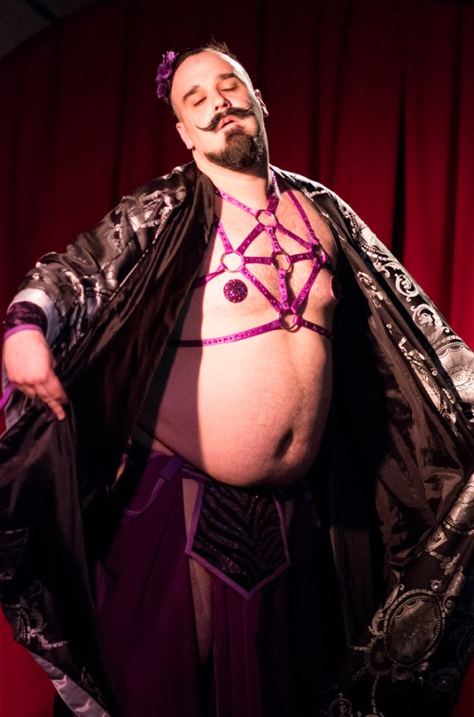 A person dressed in elaborate, feminine cosplay attire with a purple harness, lace and satin robe, and sheer purple stockings, standing confidently on a stage with red curtains in the background.