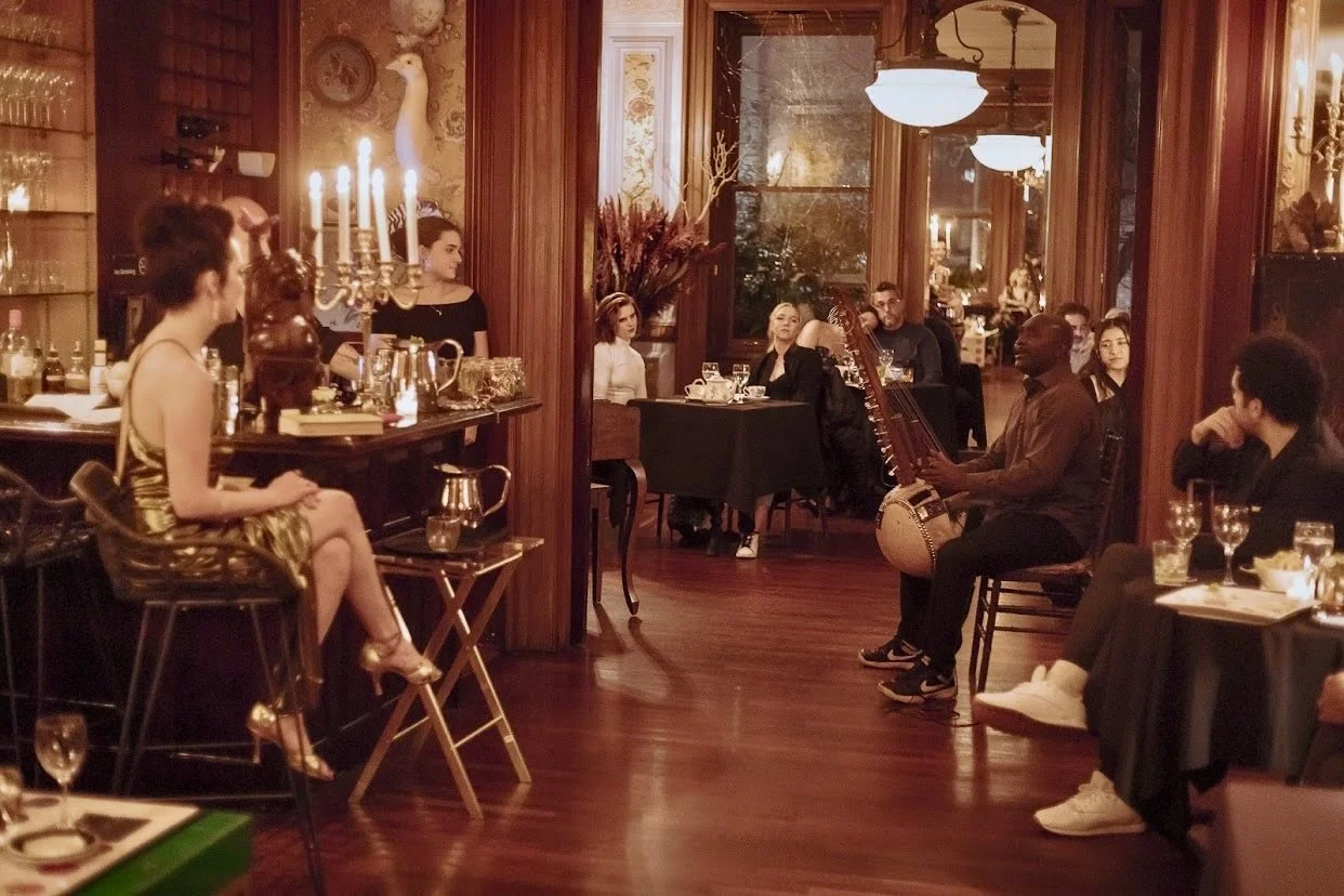 A musical performance at a cozy, elegant restaurant with a woman in a gold dress sitting on a stool, a man playing a harp, and an audience seated at tables, enjoying the show.