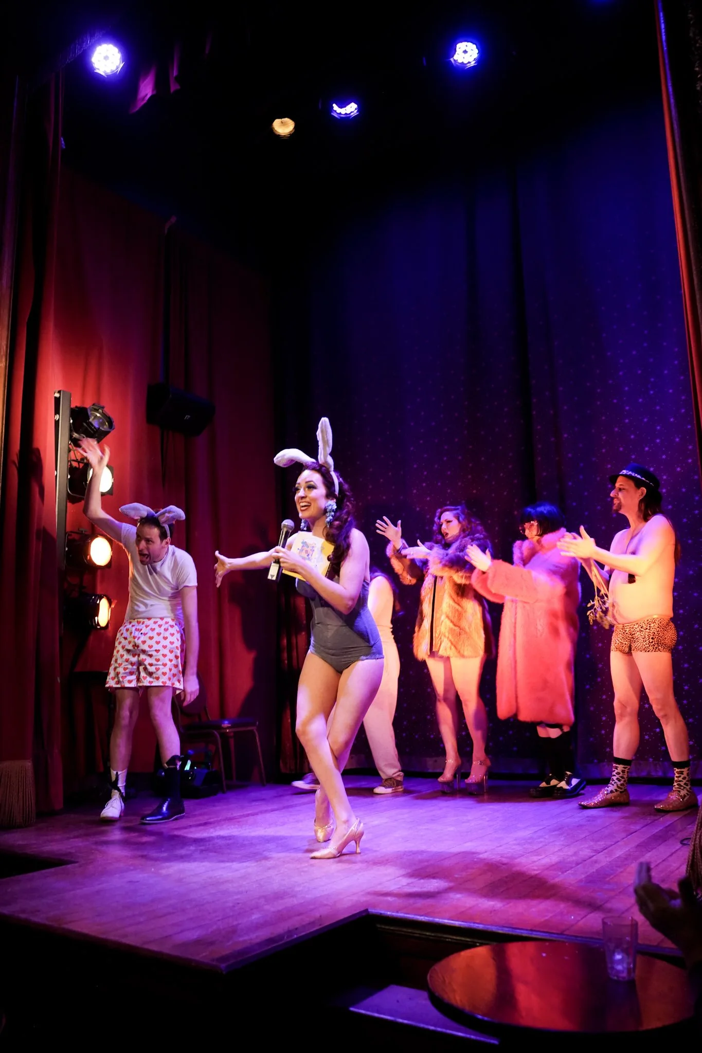 A performance on a stage with performers dressed in colorful animal and fantasy costumes, including bunny ears, a fur coat, and leopard print, under bright stage lights.