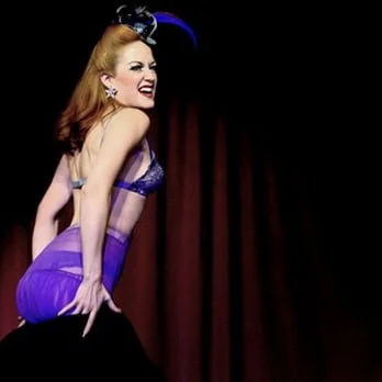 A woman with red hair and vintage-style makeup smiling and posing in front of a curtain.