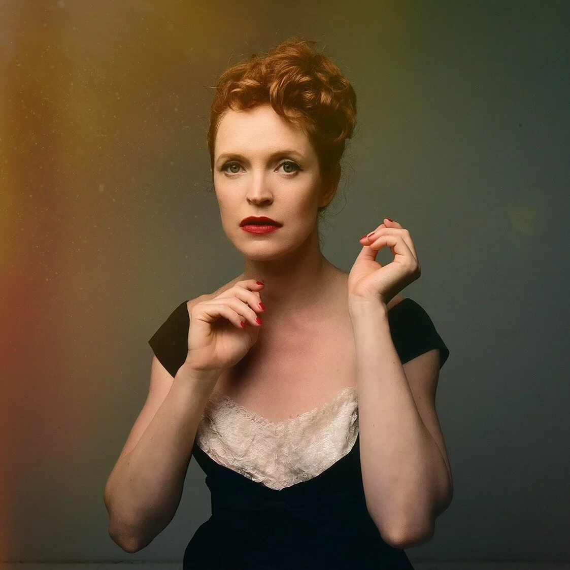A woman with red hair styled in an updo, wearing a black dress with a lace neckline, and red lipstick, standing against a neutral background.