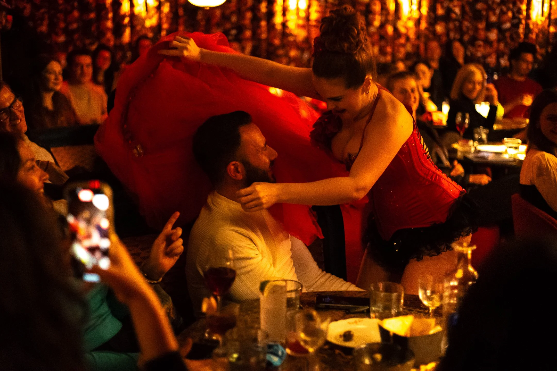 A woman in a red corset and black lace skirt dancing or performing for a man seated at a table, surrounded by seated guests in a dimly-lit setting with warm lighting and other patrons watching.