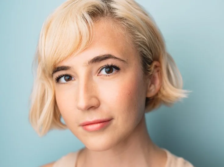 Close-up portrait of a young woman with short blonde hair and blue eyes, smiling softly, against a light blue background.