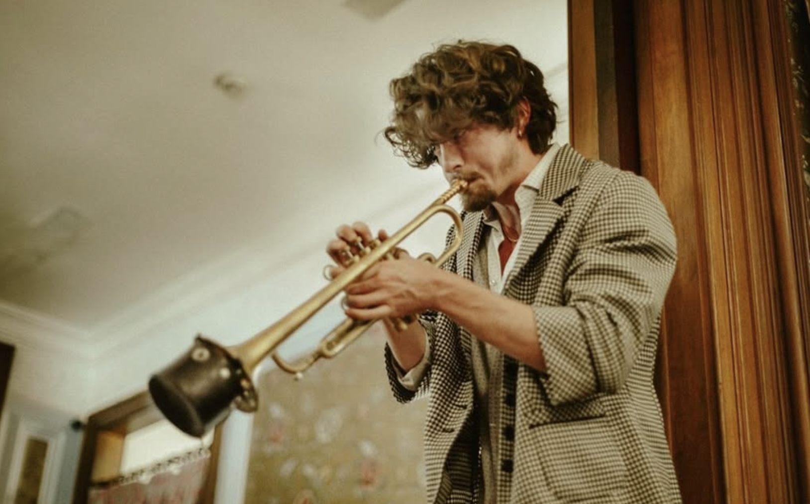 A man with curly hair plays a trumpet in a room with wooden wall paneling.