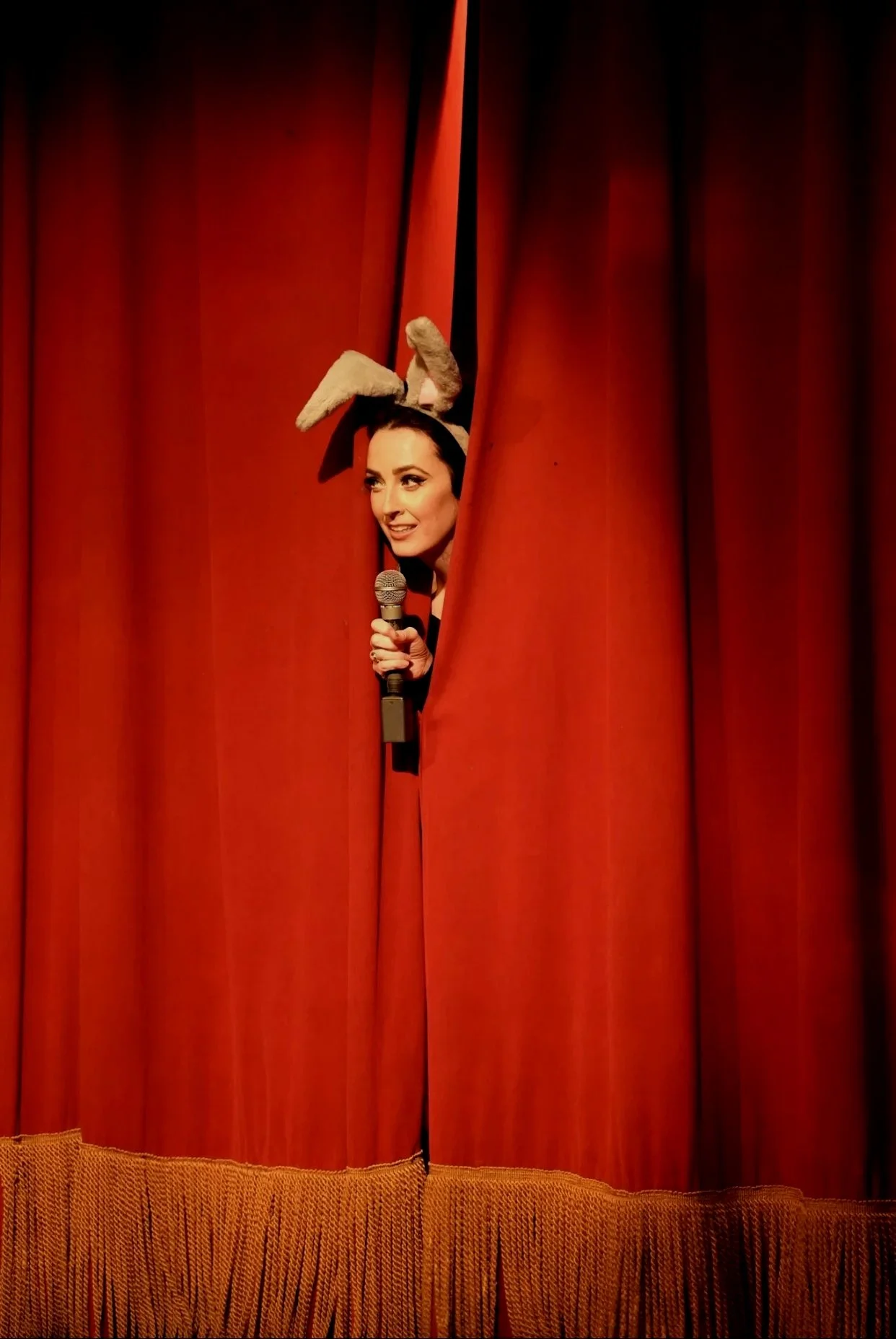 Woman with bunny ears peeking through a red theater curtain, holding a microphone and smiling.