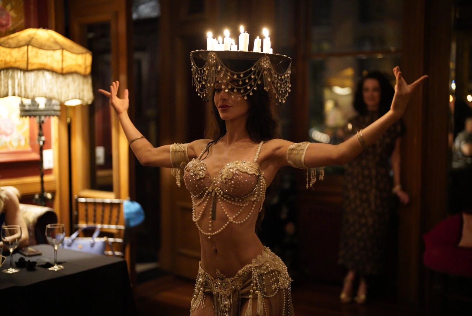 A woman wearing an ornate pearl and jewel-studded belly dance costume, with a large candlelit chandelier on her head, dancing in a dimly lit, vintage-style room.