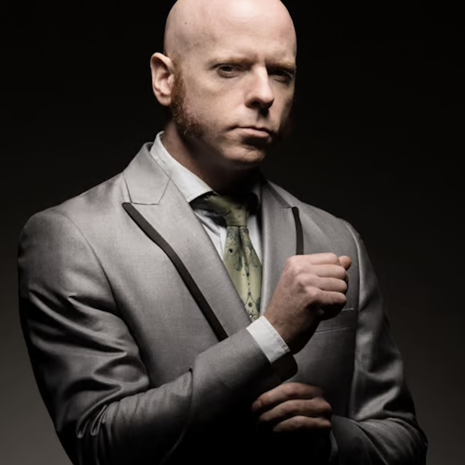 A man with a shaved head and beard in a gray suit, white shirt, and patterned green tie, posing with his glasses held in his hand against a dark background.