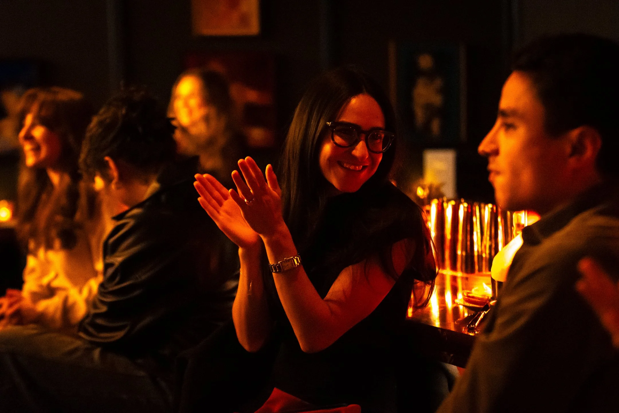 People socializing at a dimly lit bar, with a woman smiling and clapping engaged in conversation.