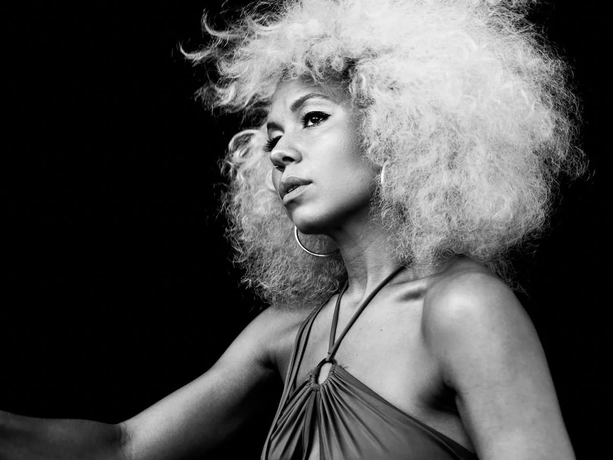 Black and white portrait of a woman with voluminous curly hair, wearing a sleeveless top and large hoop earrings, against a dark background.