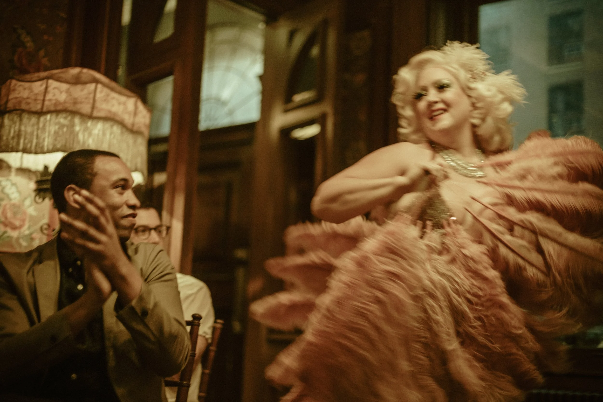 A woman with blonde curly hair in a pink dress dancing in a bar or restaurant, with her hand on her chest and smiling. A man with dark hair and a beige jacket sits at the table, watching her dance, with his hand near his face.