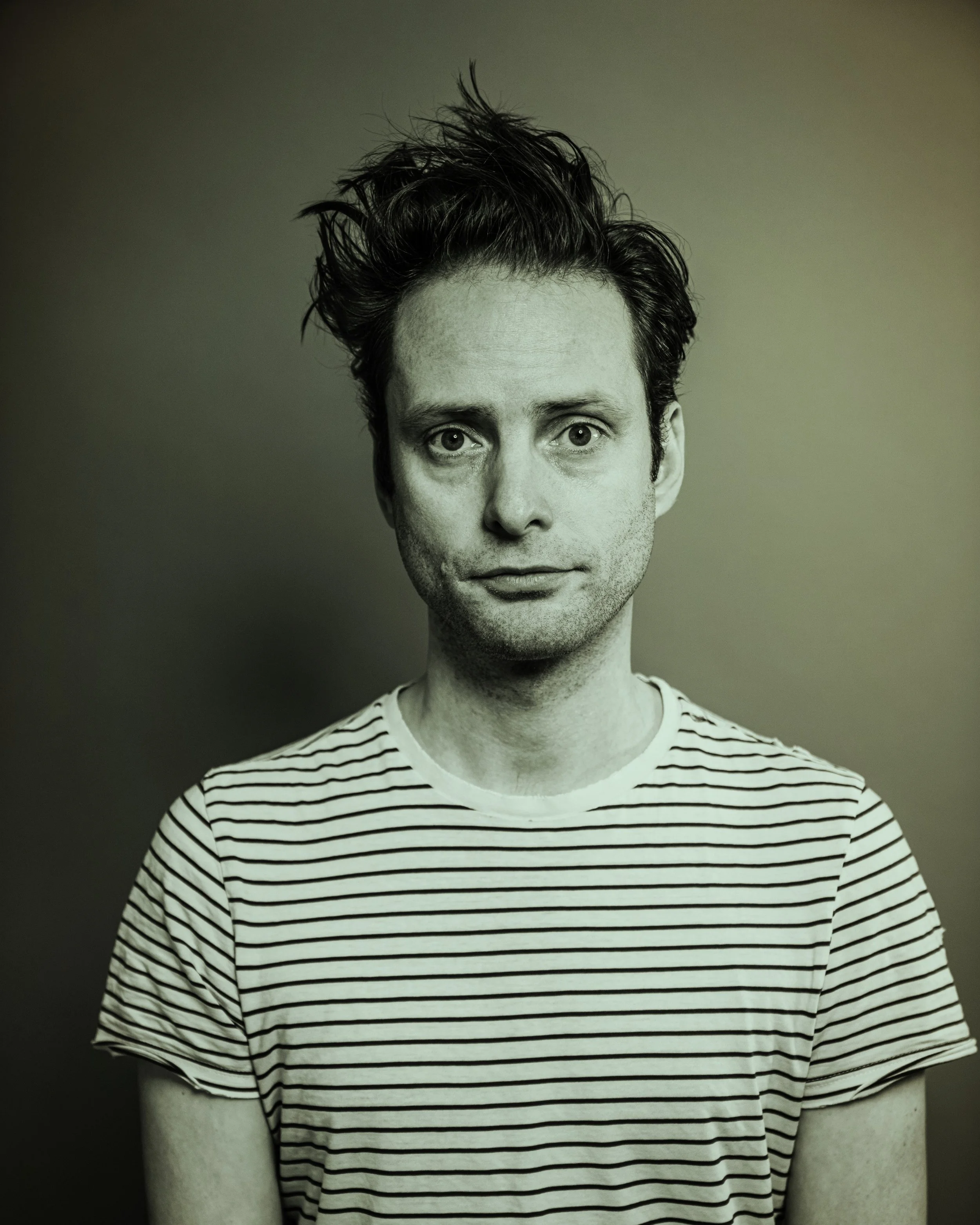 Black and white portrait of a man with messy hair wearing a striped t-shirt, looking directly at the camera.