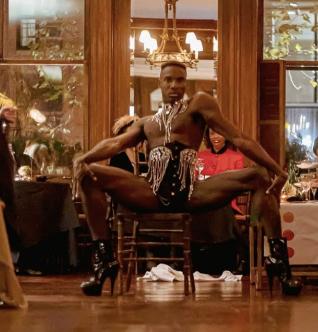 A man dressed in black with silver embellishments poses in a split position on a wooden chair at a party or event. He is wearing high-heeled boots and has a confident expression, with people and a chandelier visible in the background.