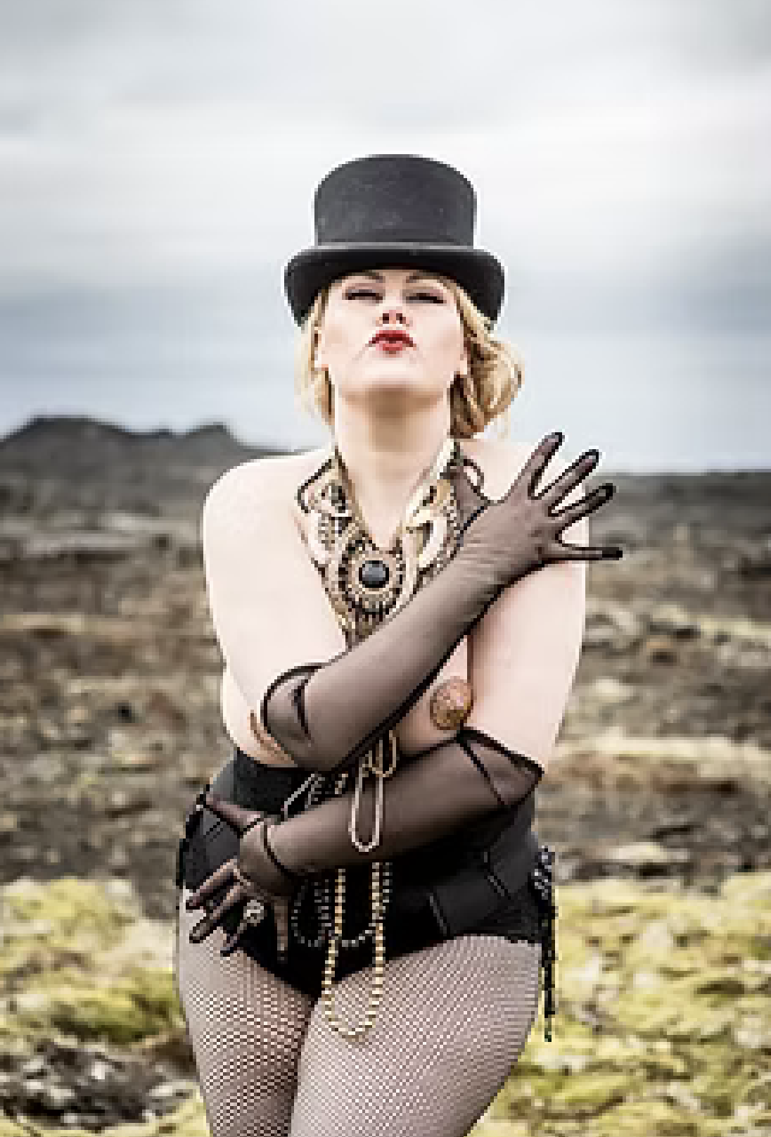 A woman standing outdoors in a rugged landscape, wearing a top hat, leopard-print jewelry, sheer black gloves, fishnet stockings, and a black leather skirt.