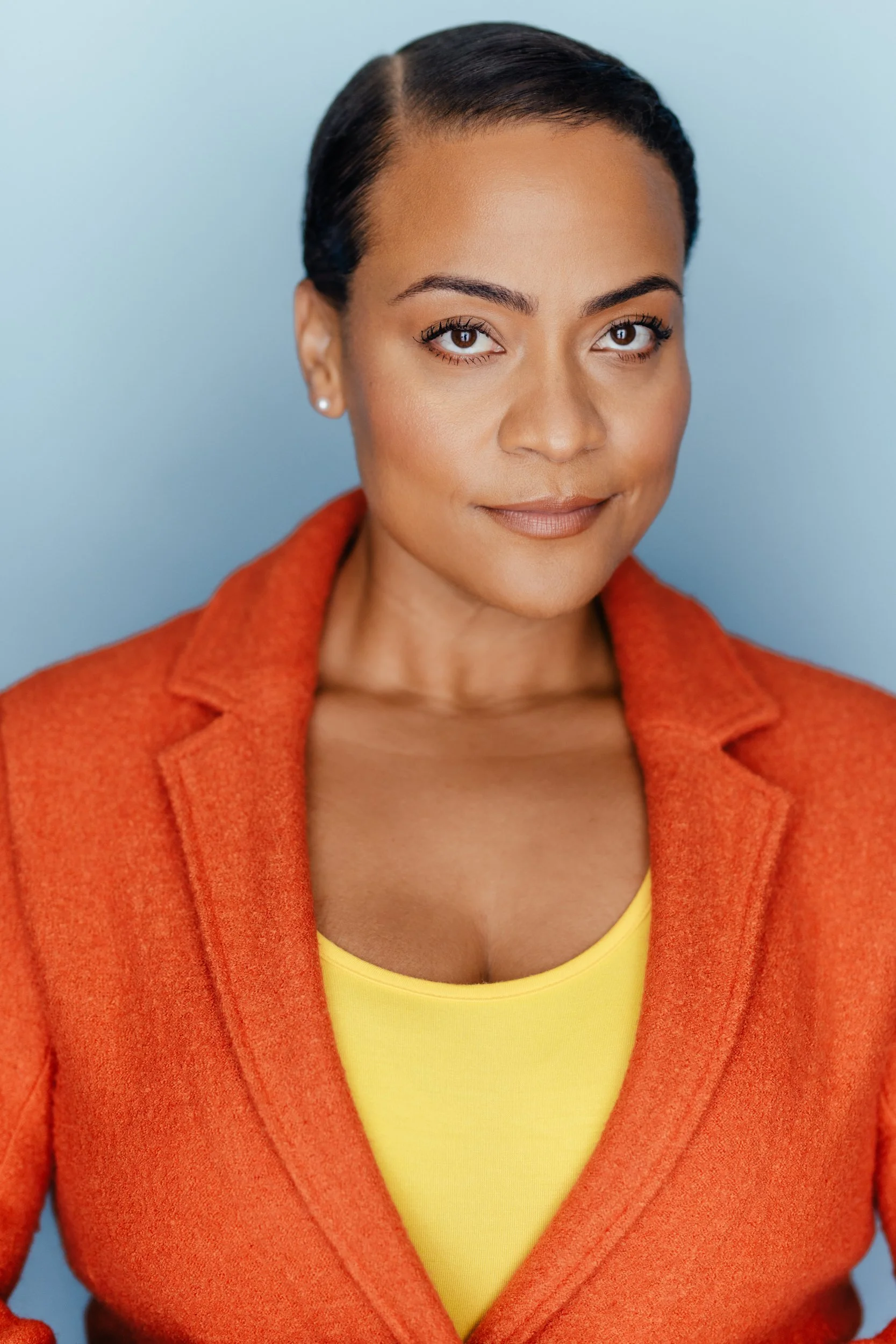 A woman with short black hair, wearing an orange blazer over a yellow top, standing against a light blue background.