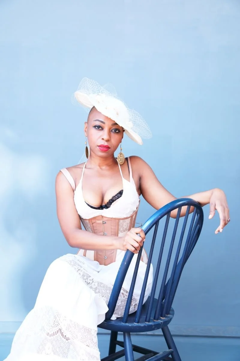A woman in vintage-style clothing and a large white hat, sitting on a blue chair against a plain blue background.