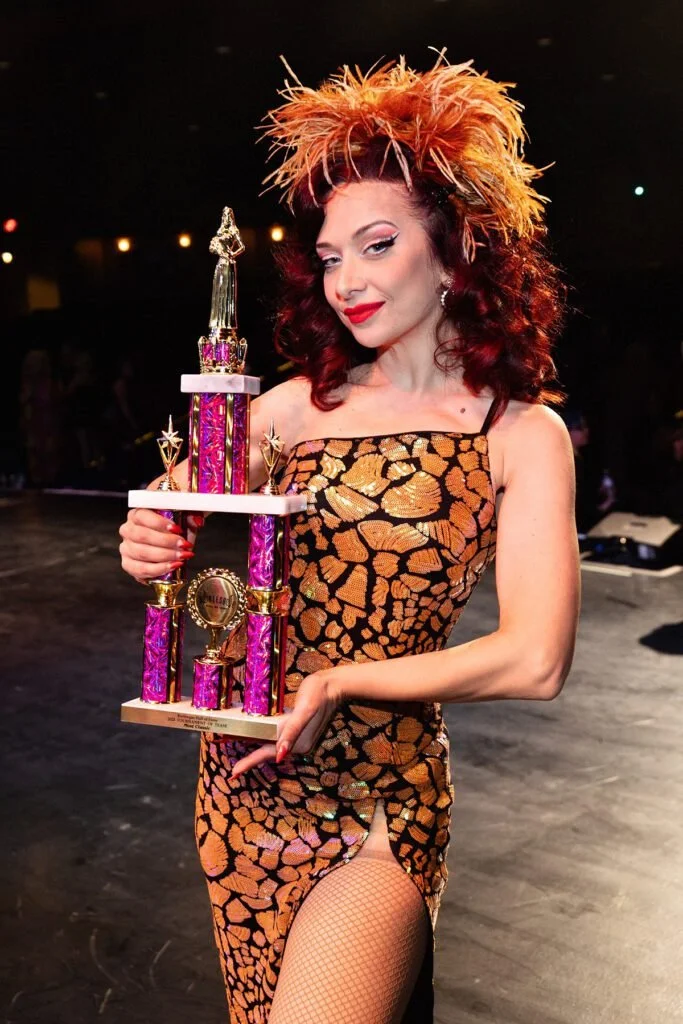 A woman in a leopard print dress with fishnet stockings holding a large colorful trophy while standing on a stage or dance floor.