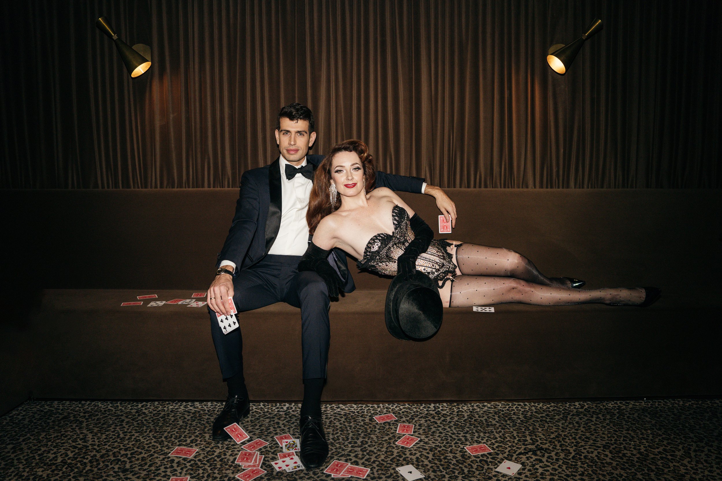 A man in a tuxedo and a woman in a glamorous black dress and gloves are sitting on a brown couch with scattered playing cards on the floor and couch. The woman is holding a black hat and the man has his arm around her. The background features dark curtains and two wall-mounted lights.