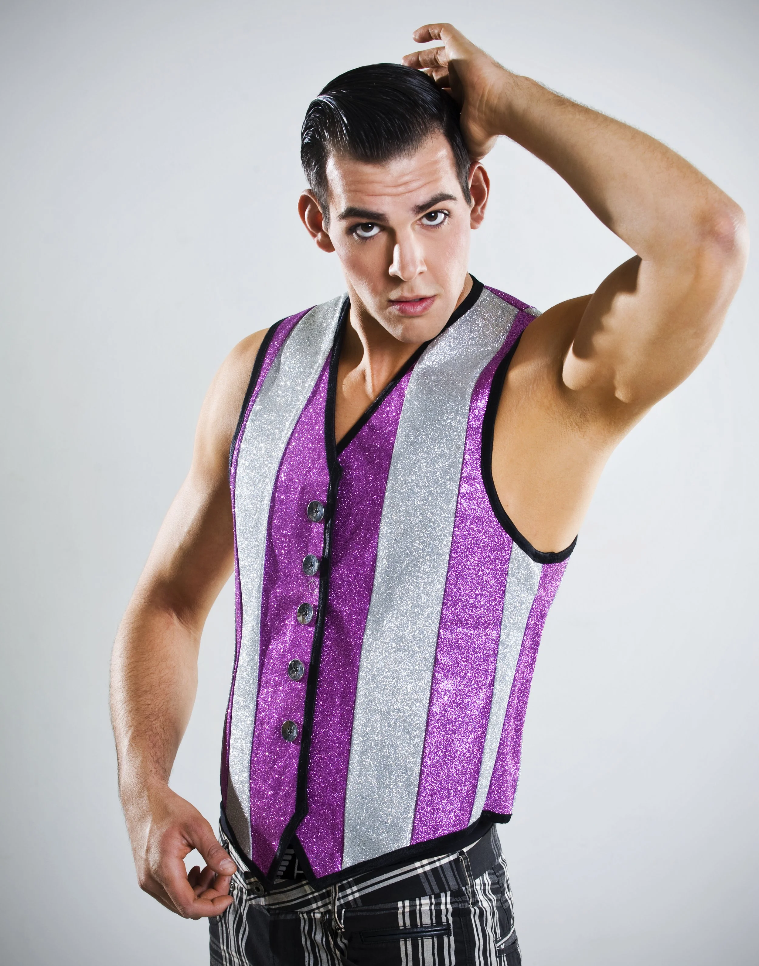 A young man with dark hair styled back, wearing a sleeveless purple and silver glitter vest, with one hand on his head, looking into the camera, against a light gray background.