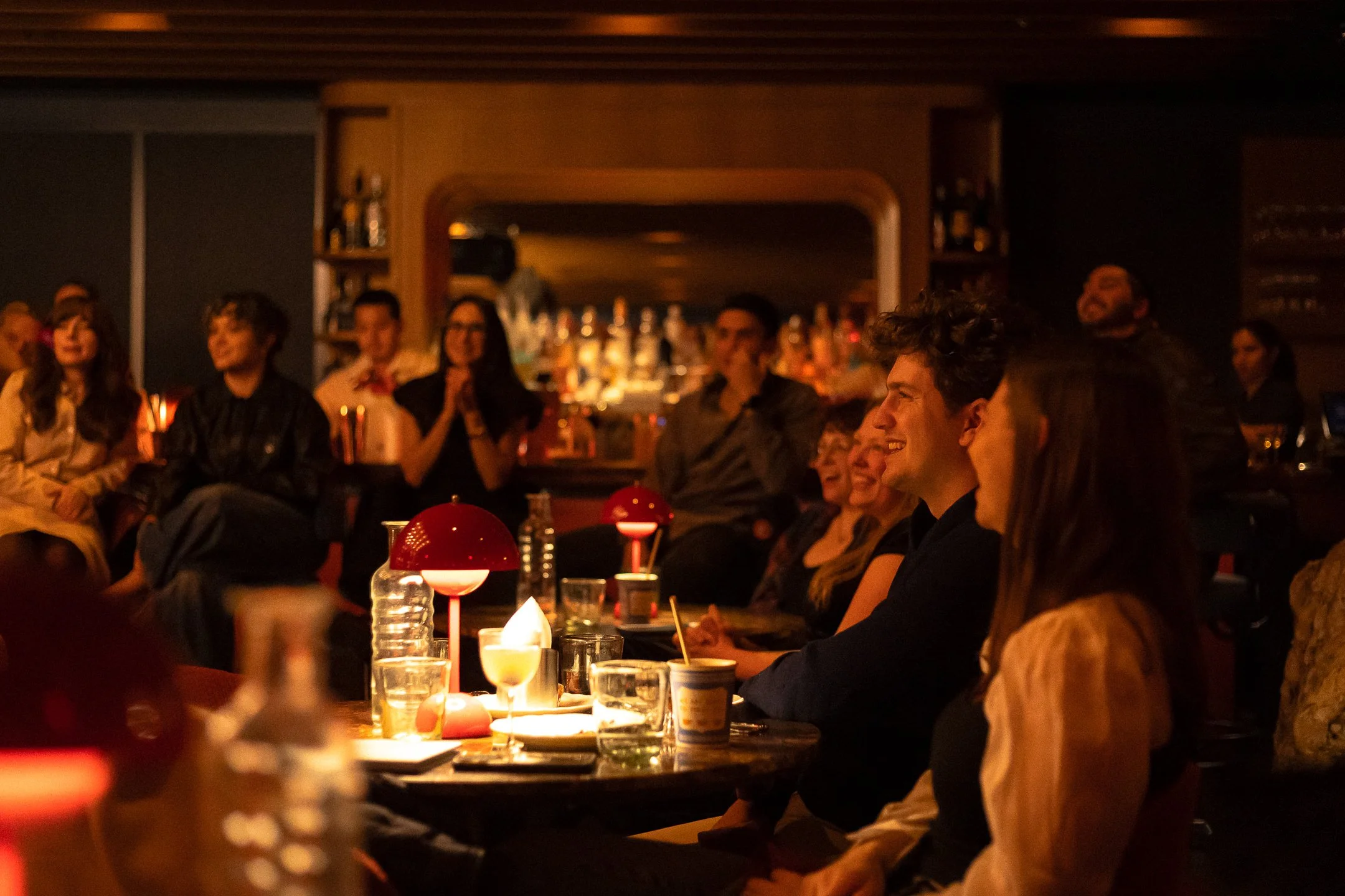 People sitting and watching entertainment at a dimly lit bar or lounge with warm lighting, bottles of alcohol on shelves, and glasses on tables.