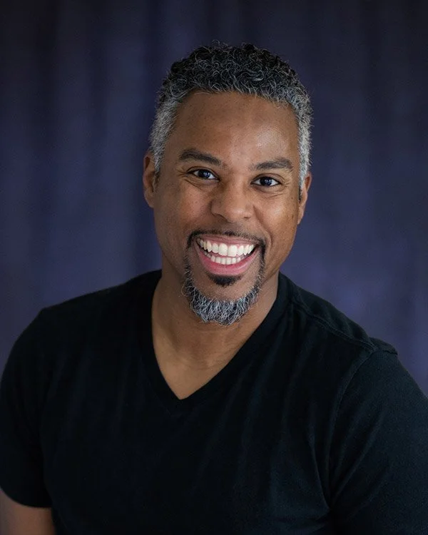 Smiling man with short curly gray hair and beard wearing a black t-shirt against a dark purple background.