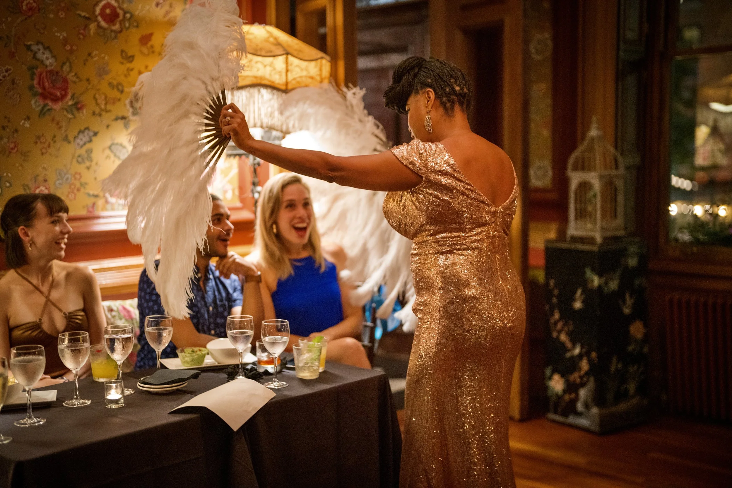 A woman in a glittering gold dress holding large white feathered fan in front of three seated friends at a dinner table in a warmly lit, decorated restaurant.