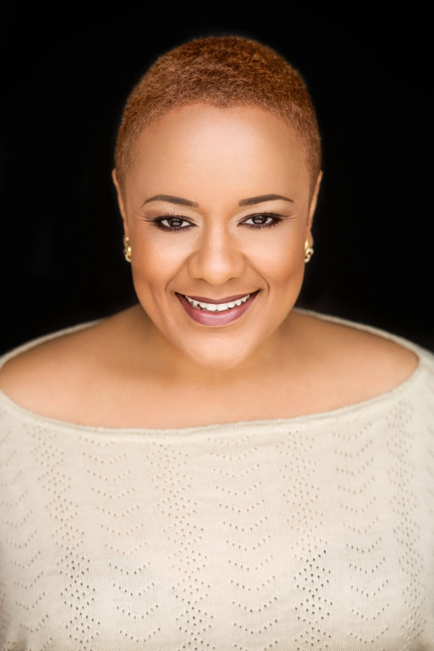 Smiling woman with short, reddish hair, wearing a light-colored knitted top and earrings, against a black background.