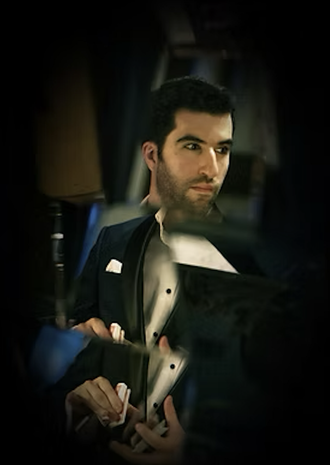 A man with dark hair, dressed in formal attire, looking intently at something off-camera, reflected in a mirror or glass surface.