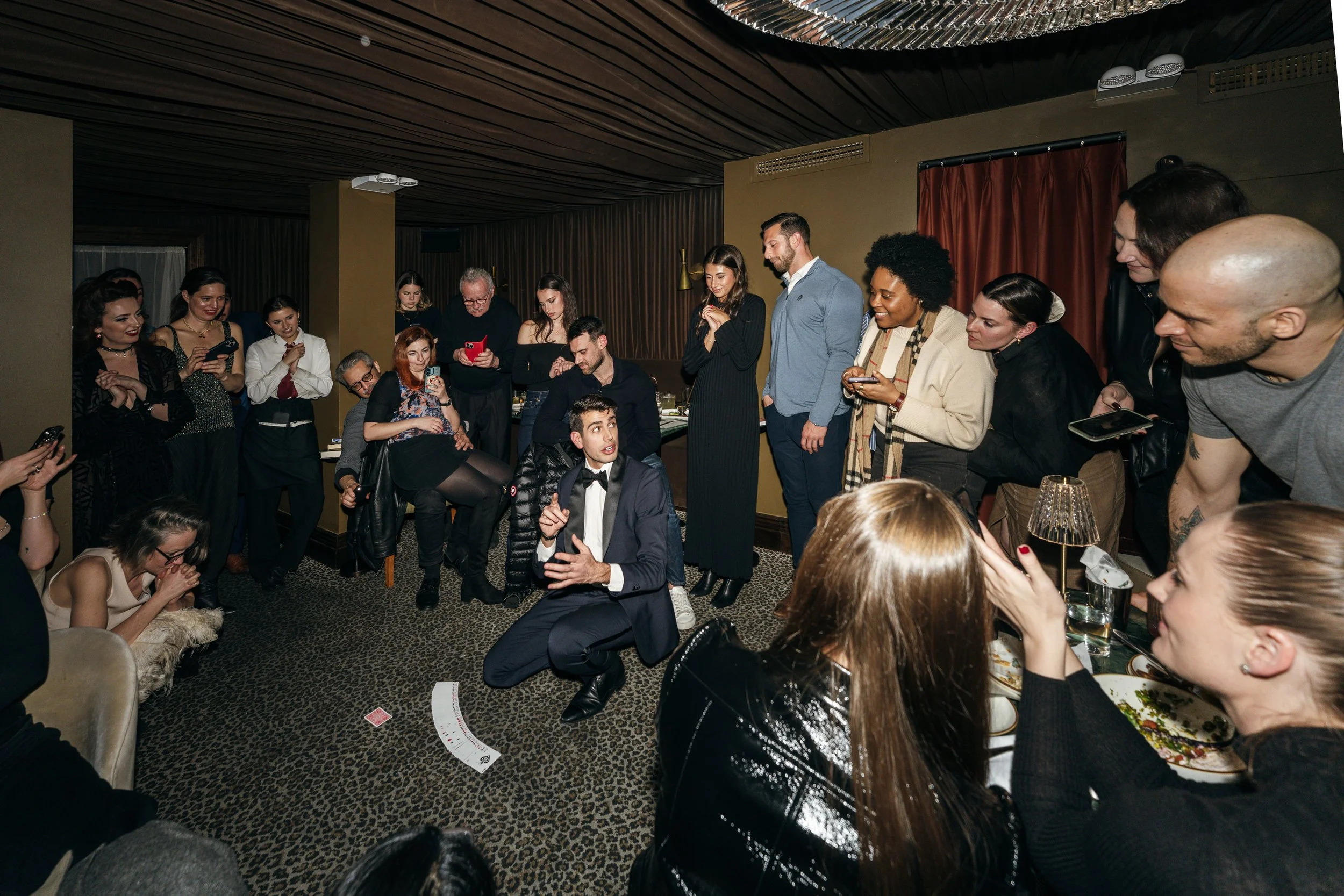 A group of people gathered in a circle around a man in a tuxedo who is kneeling and talking, at a social event in a decorated room with animal print carpet.