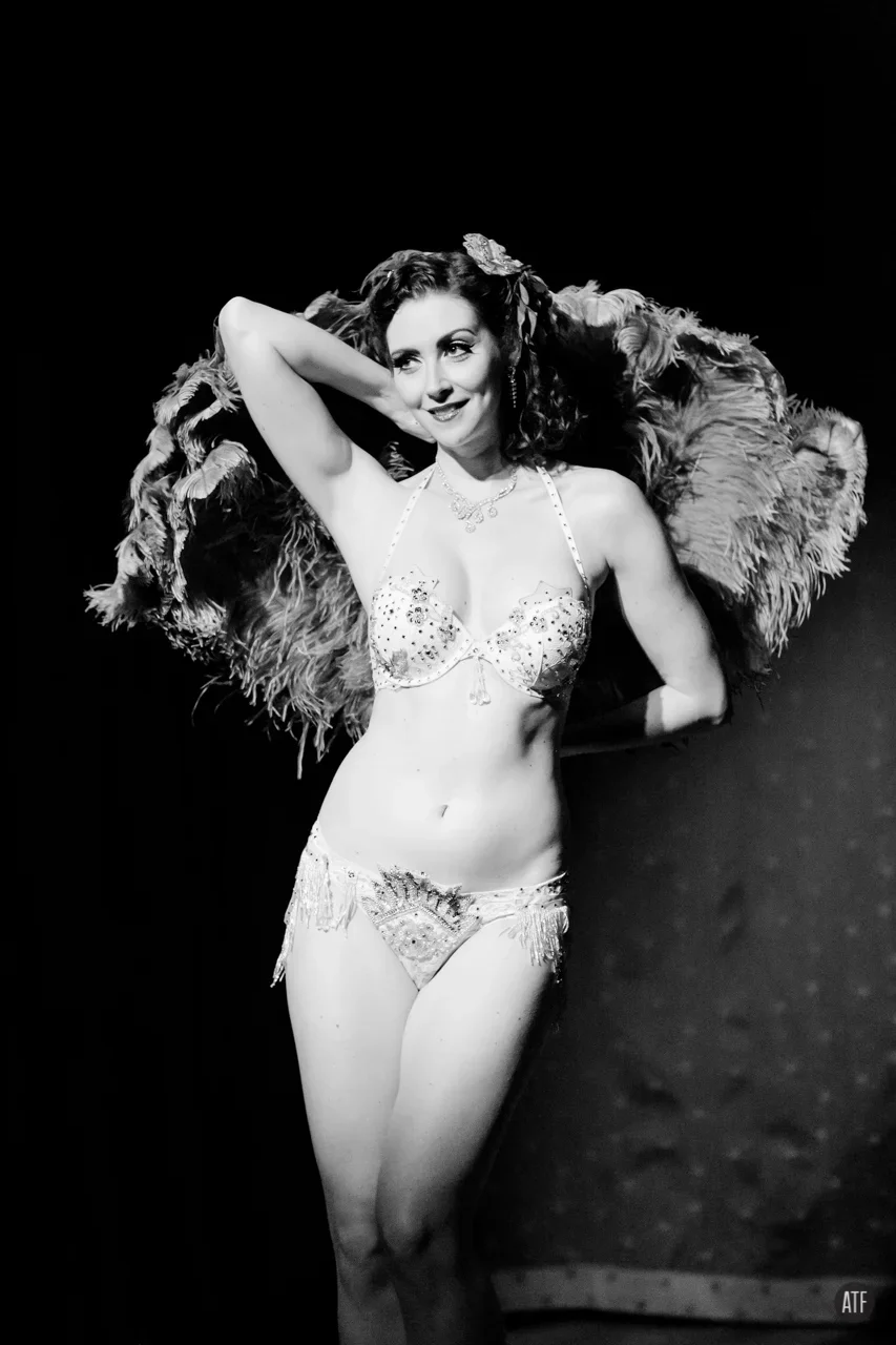 A woman in vintage lingerie poses on stage with feathered accessories, smiling and striking a confident pose in a black and white photo.
